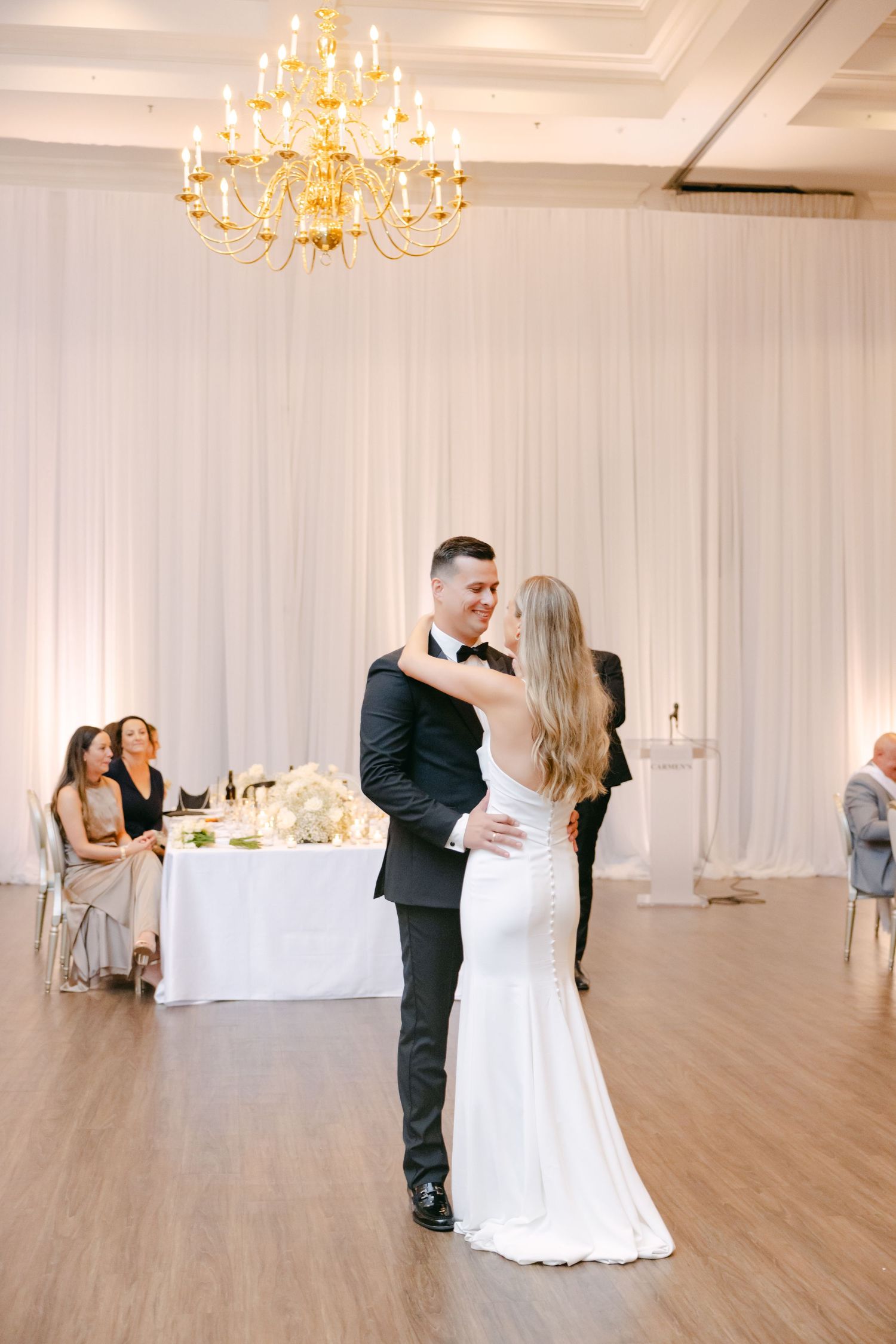 Elegant first dance under golden chandelier with white draping and reception tables in background.