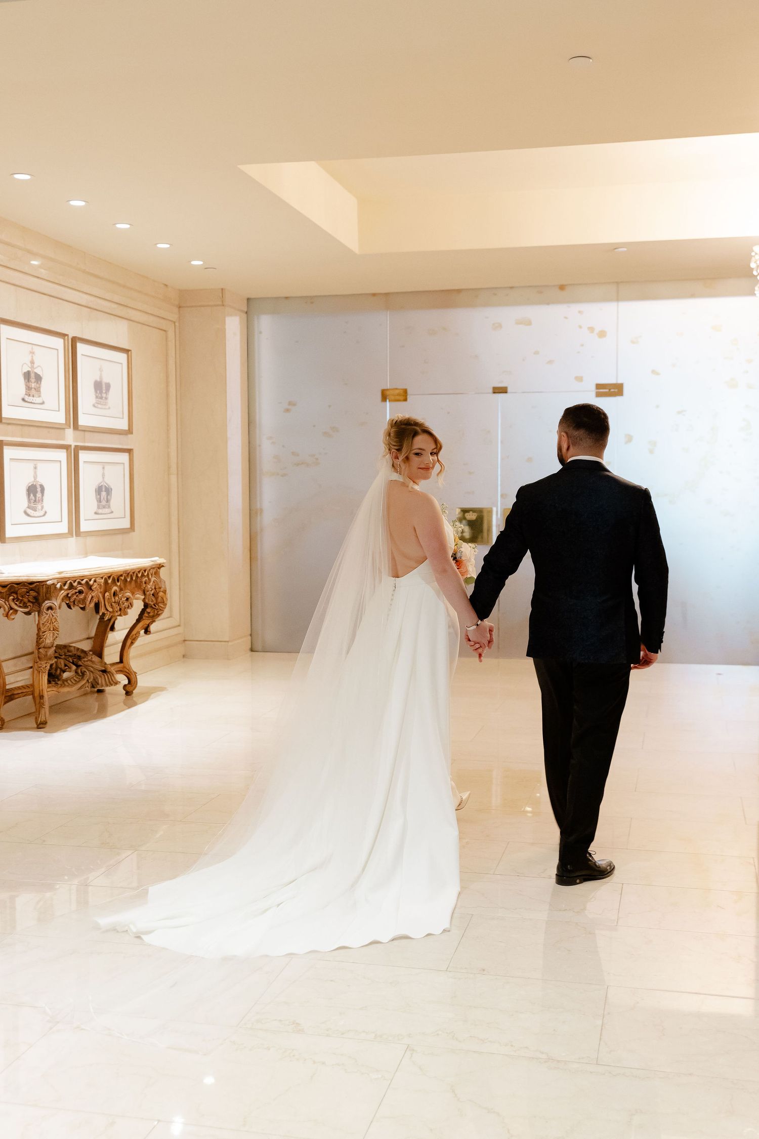Bride and groom walking through an elegant white hallway with a long flowing wedding dress train.