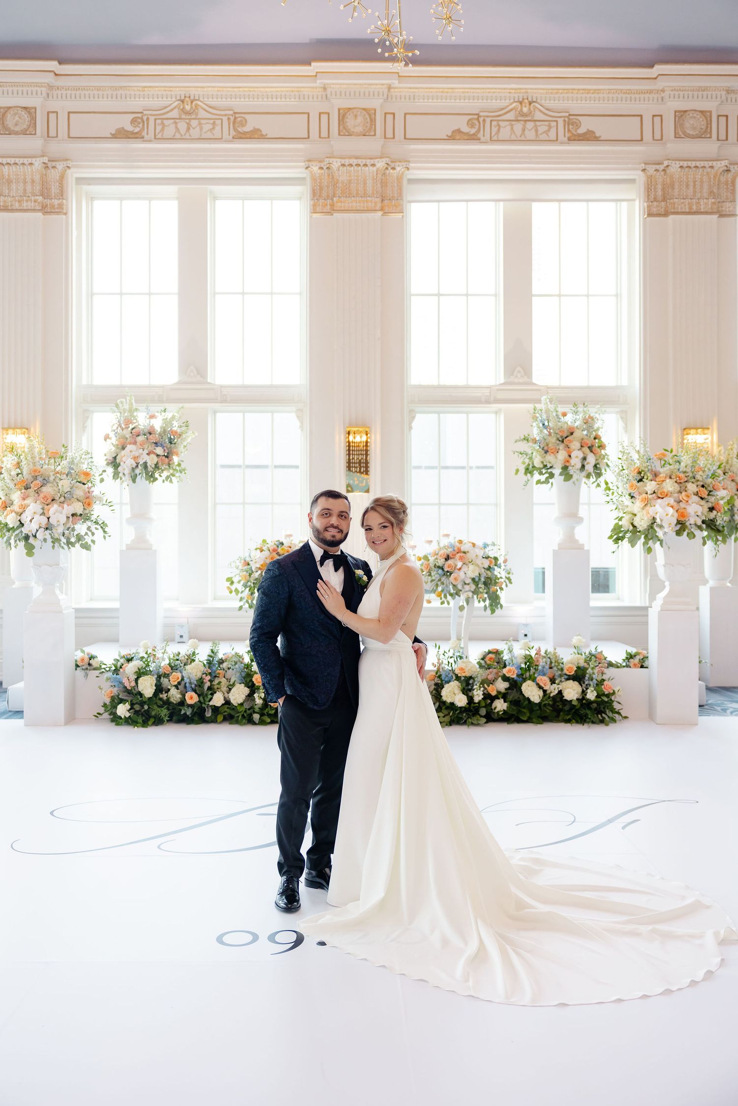 Elegant wedding ceremony setup in white room with tall floral arrangements and natural light streaming through large windows.