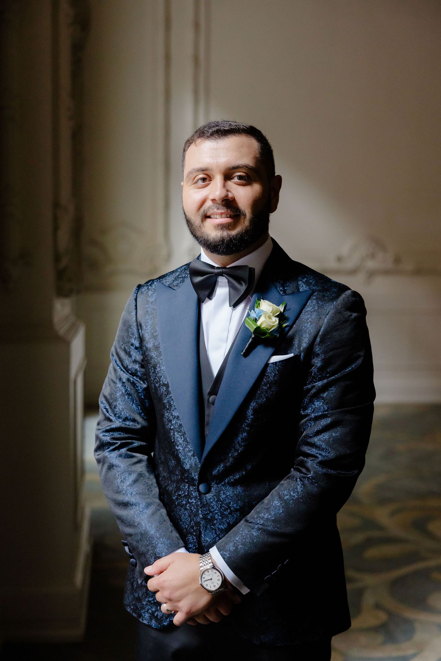 Groom poses in a textured navy blue tuxedo jacket with boutonniere against neutral background.