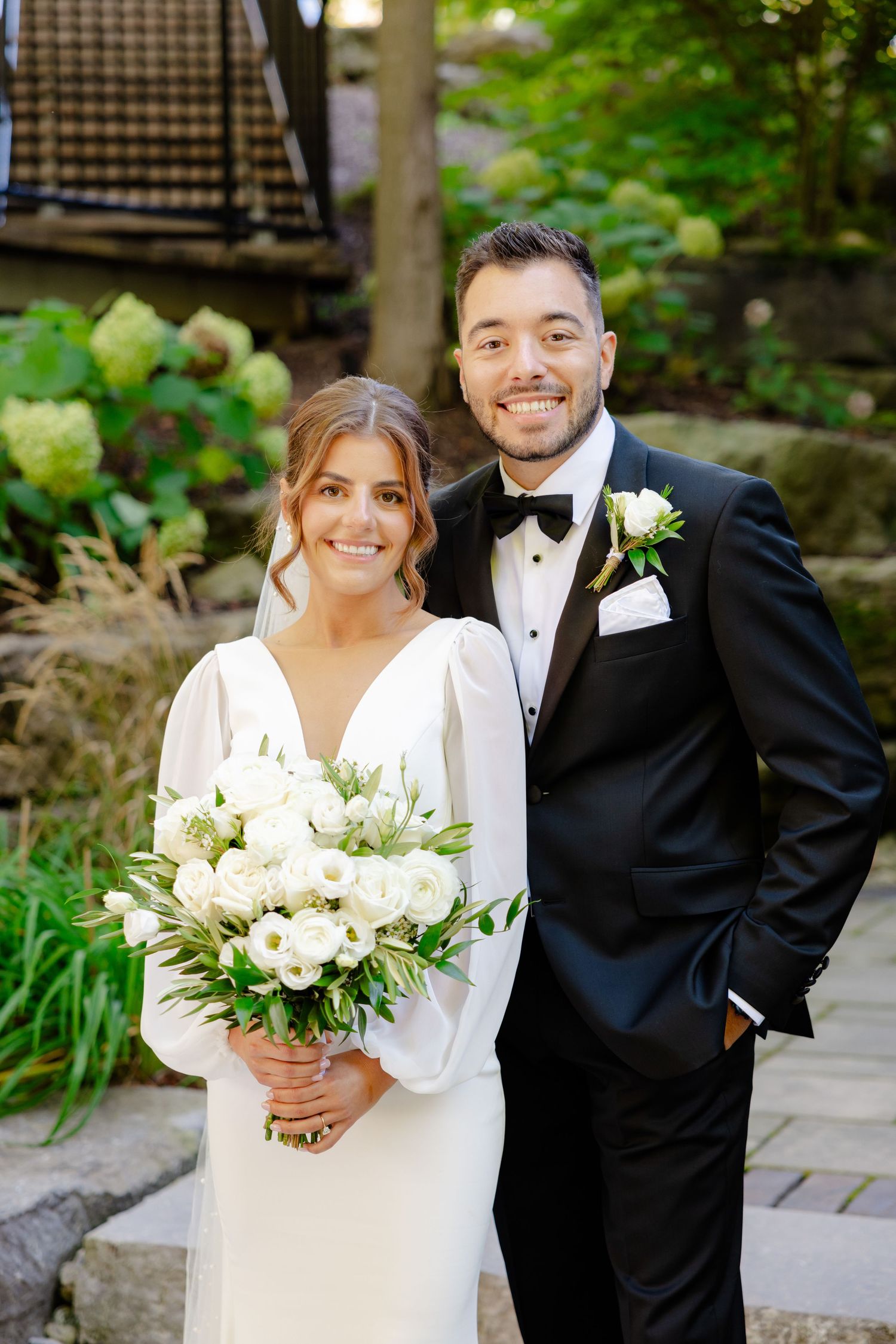 Bride in elegant white dress holds bouquet while posing with groom in black tuxedo against garden backdrop.