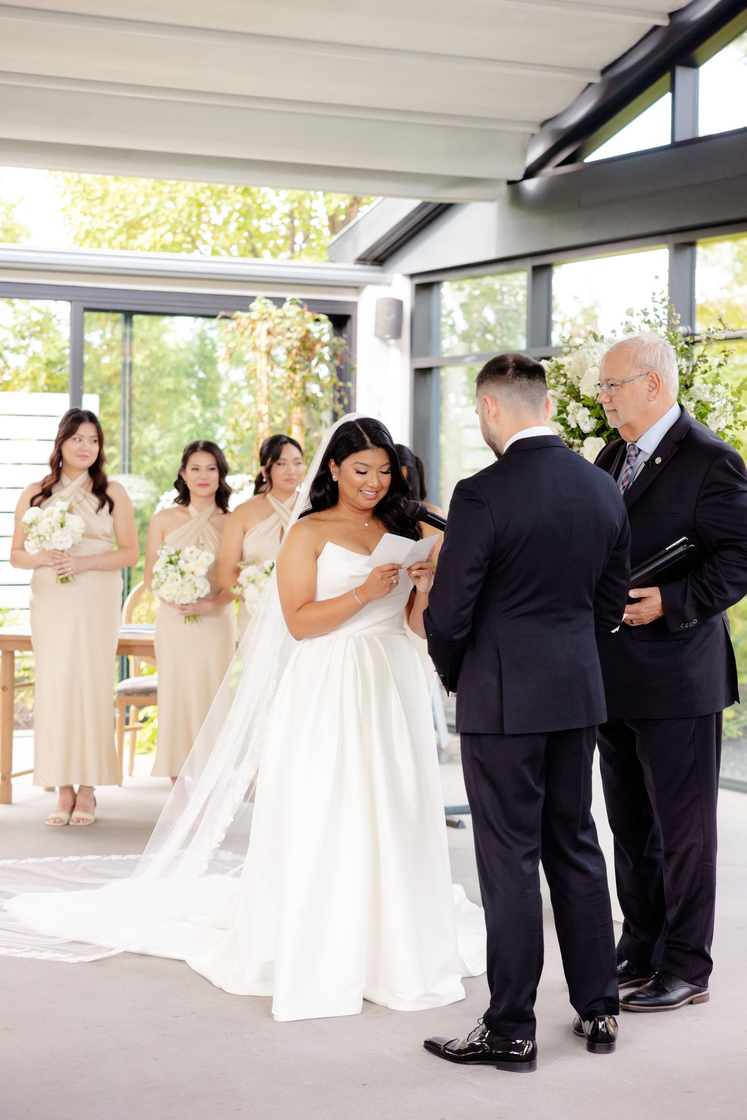 Indoor wedding ceremony with bridesmaids in blush dresses and white floral bouquets.
