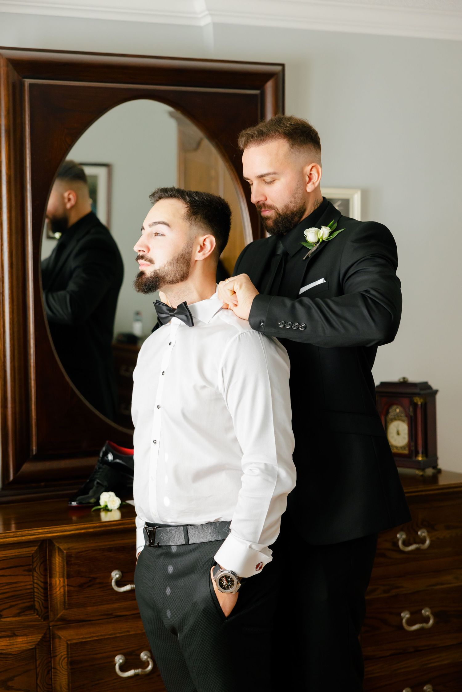 Two people in formal wear making final adjustments to attire in front of an ornate mirror.