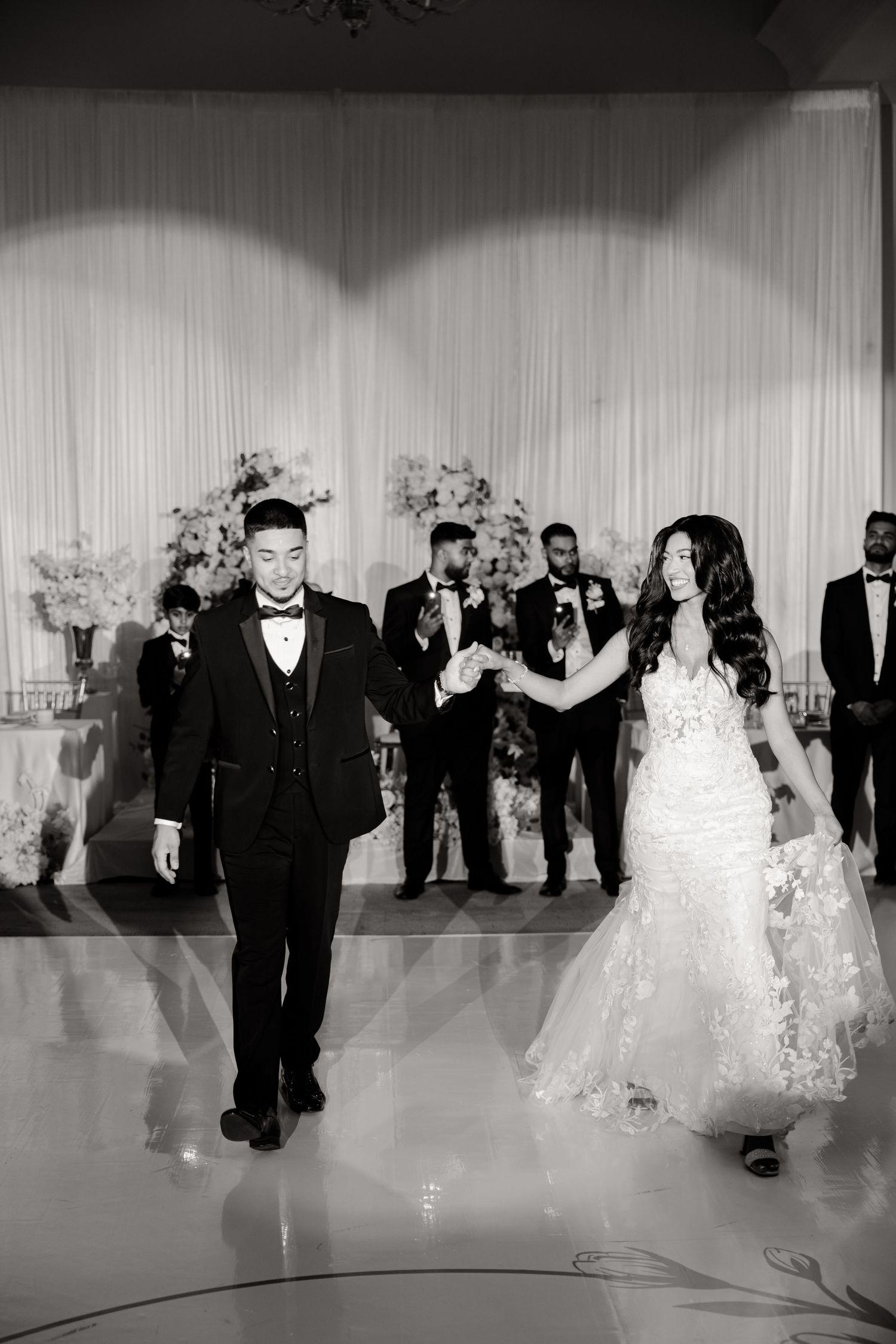 A newly married couple walks together at their wedding reception with guests and floral decor in the background.