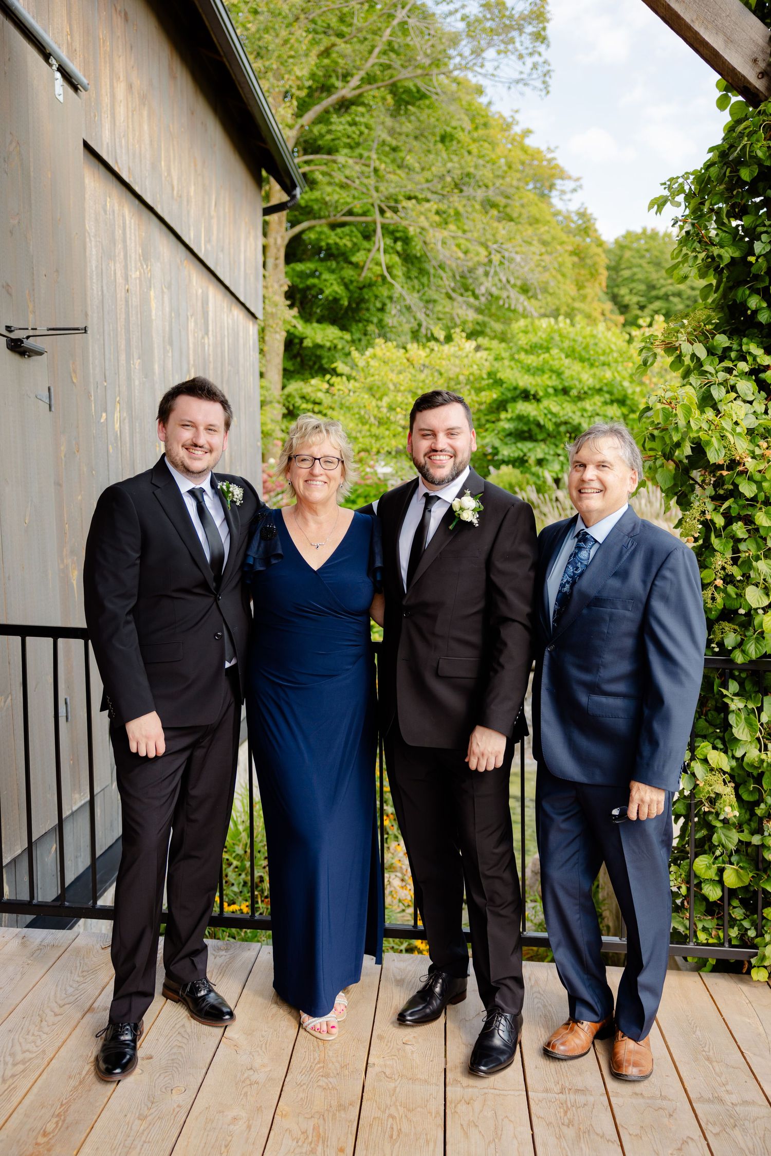 Four people in formal attire pose on wooden deck in garden setting.