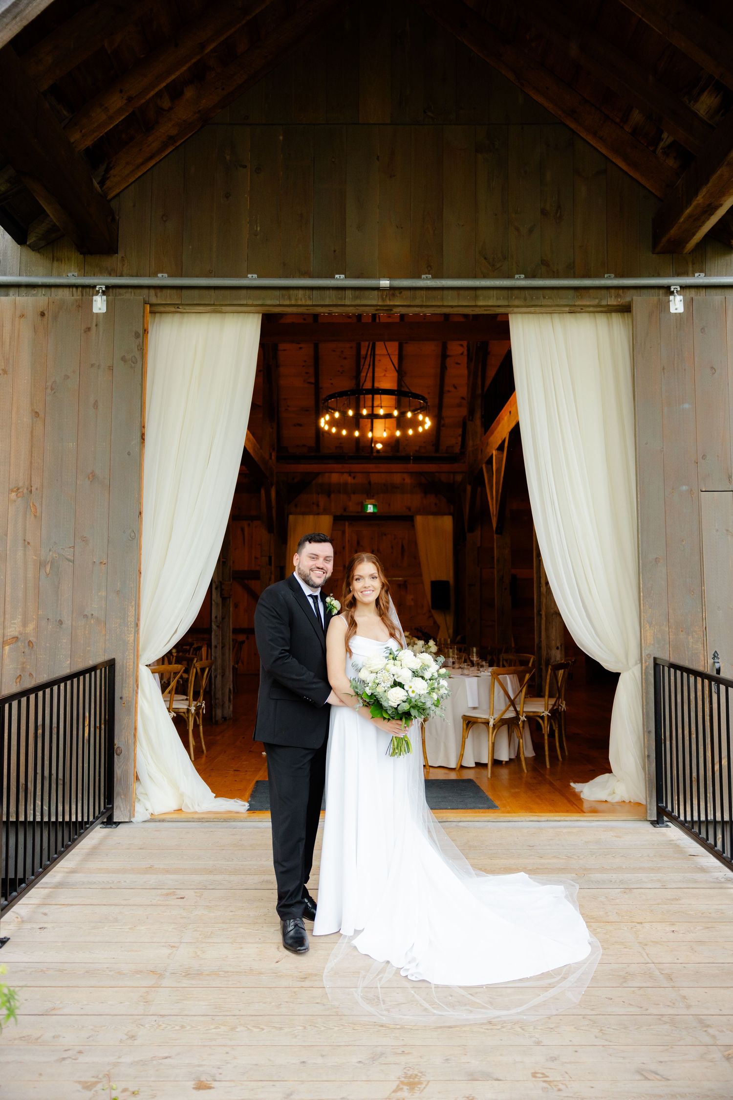 Wedding couple poses at rustic barn venue entrance with flowing white curtains and chandelier.