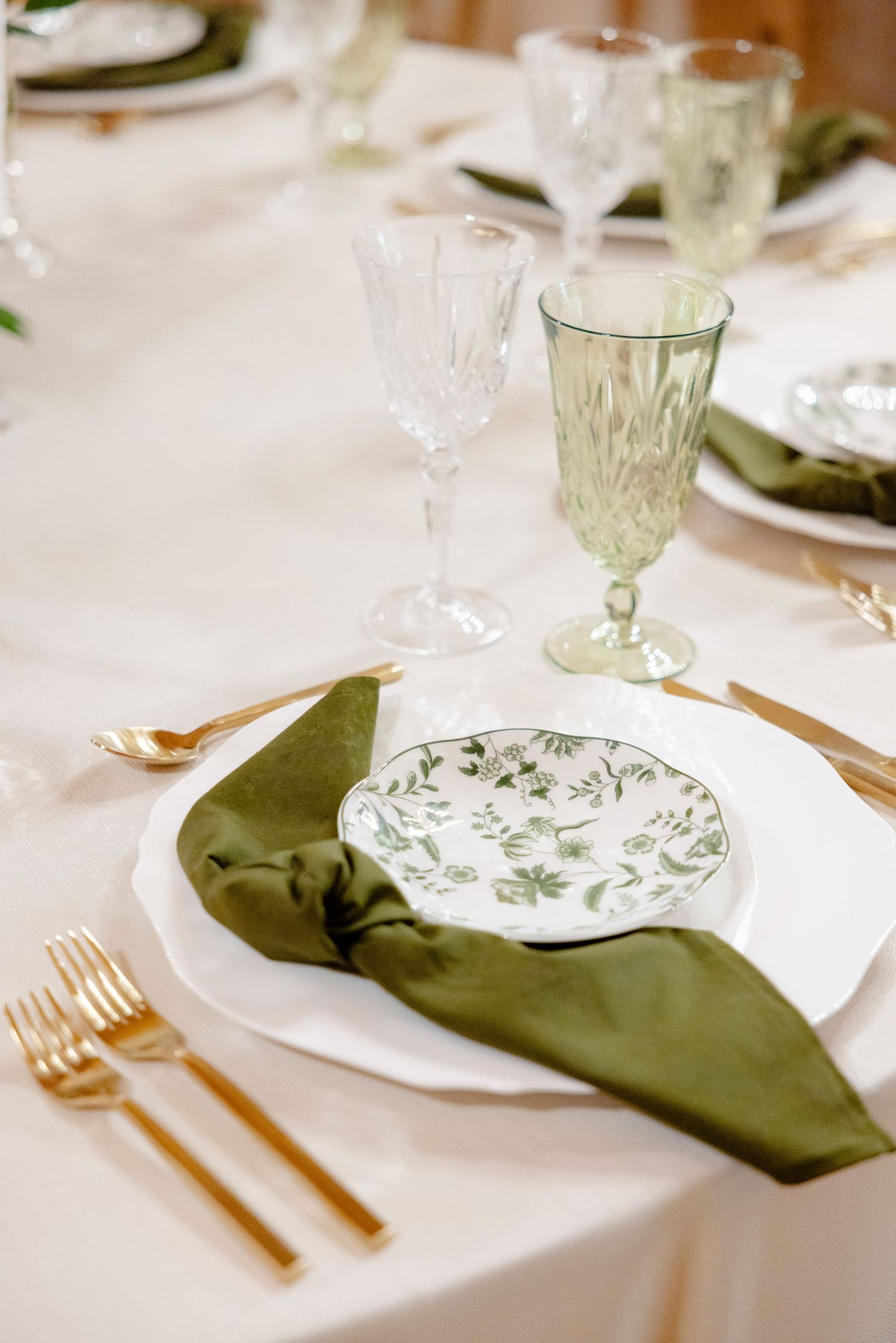Vintage-style place setting with green napkin, gold flatware, and patterned plate on white tablecloth.