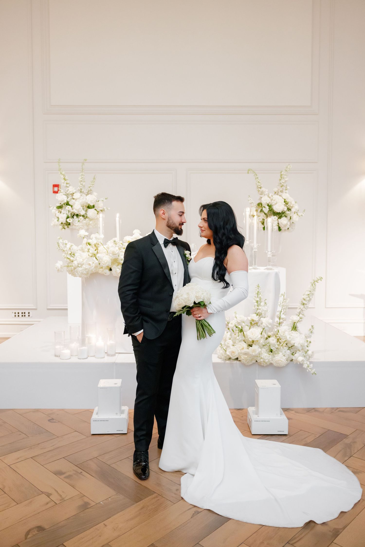 Wedding couple shares intimate moment against white wall with elegant floral decor.