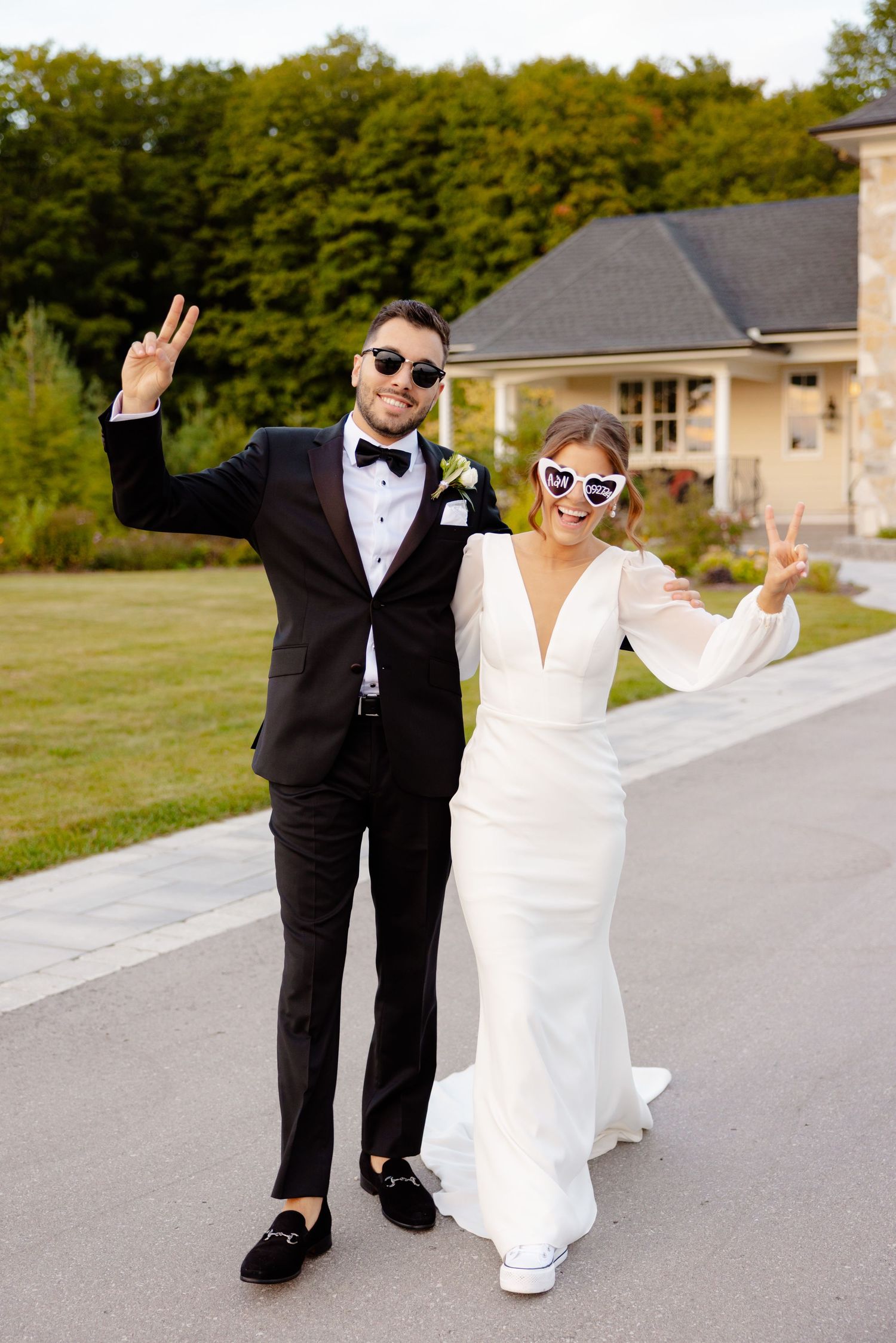 A celebratory wedding couple in formal attire makes peace signs while wearing sunglasses.