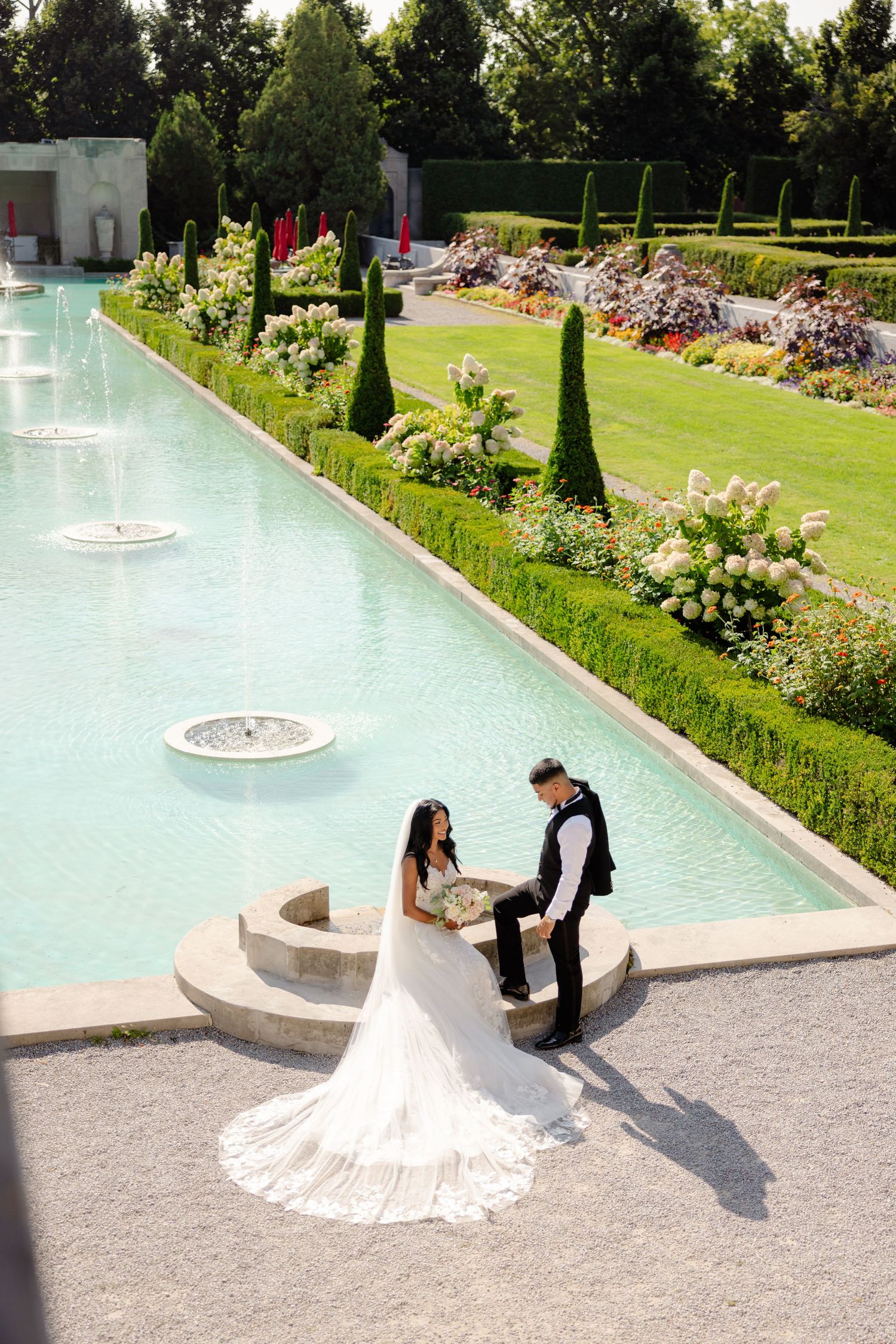 A wedding couple shares a moment beside a reflective pool lined with cypress trees and blooming flowers.