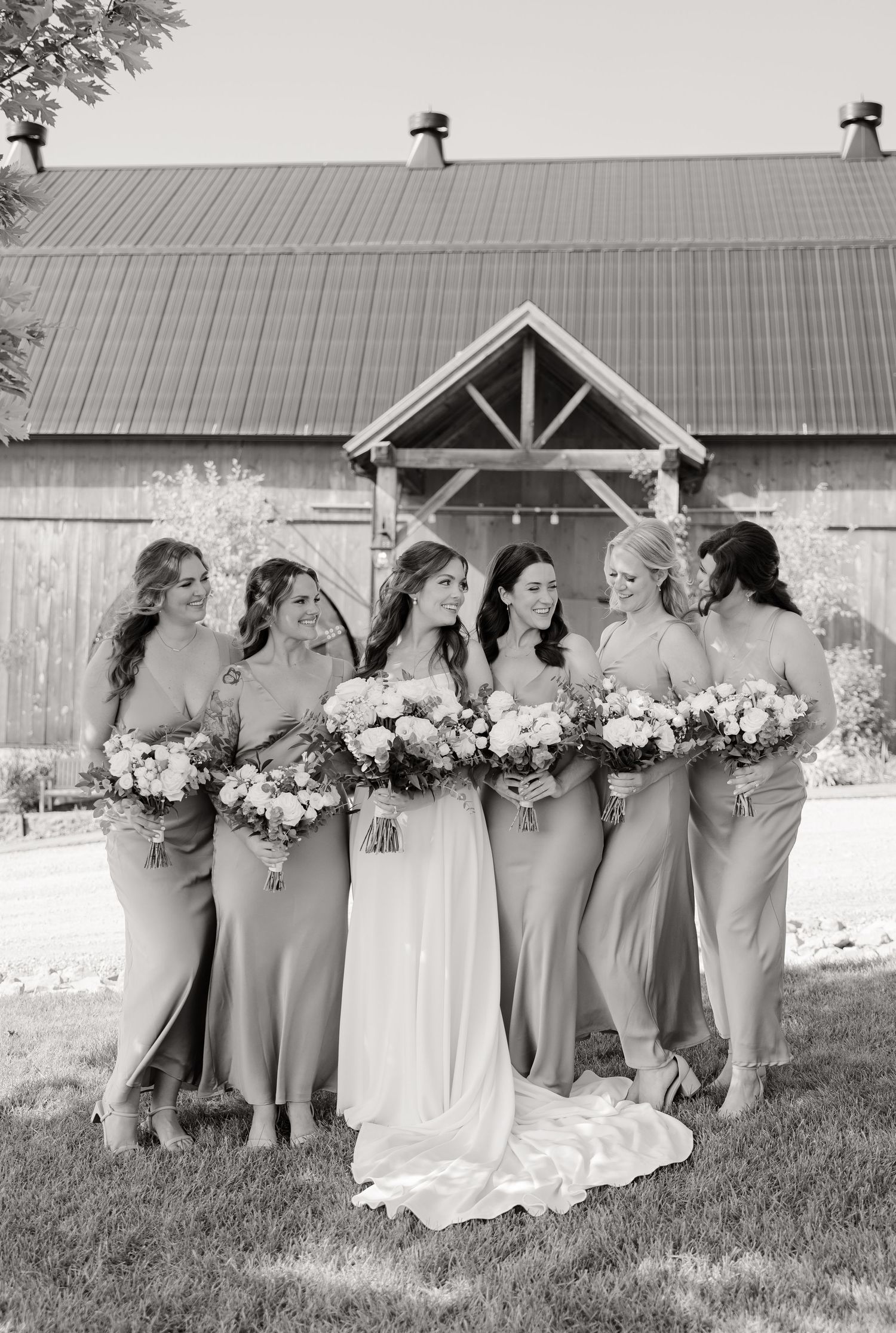 Black and white portrait of bridal party with elegant bouquets in front of barn venue.