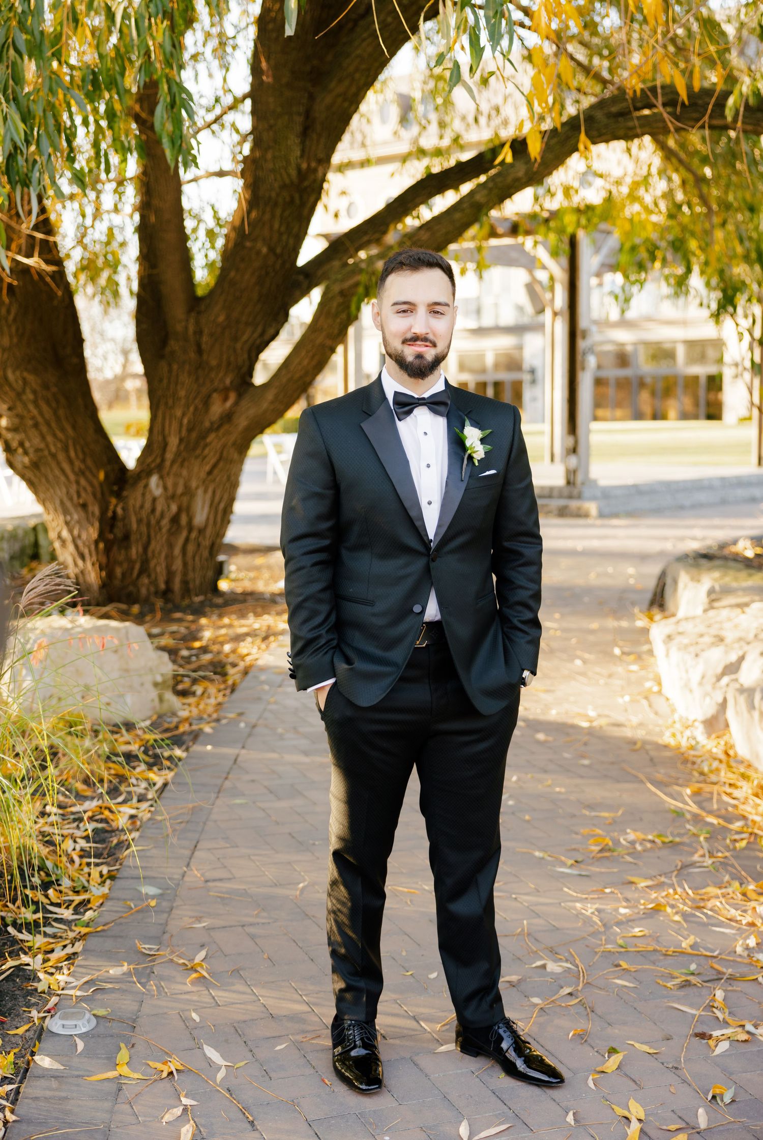 Groom in classic black tuxedo standing on stone path under willow tree.
