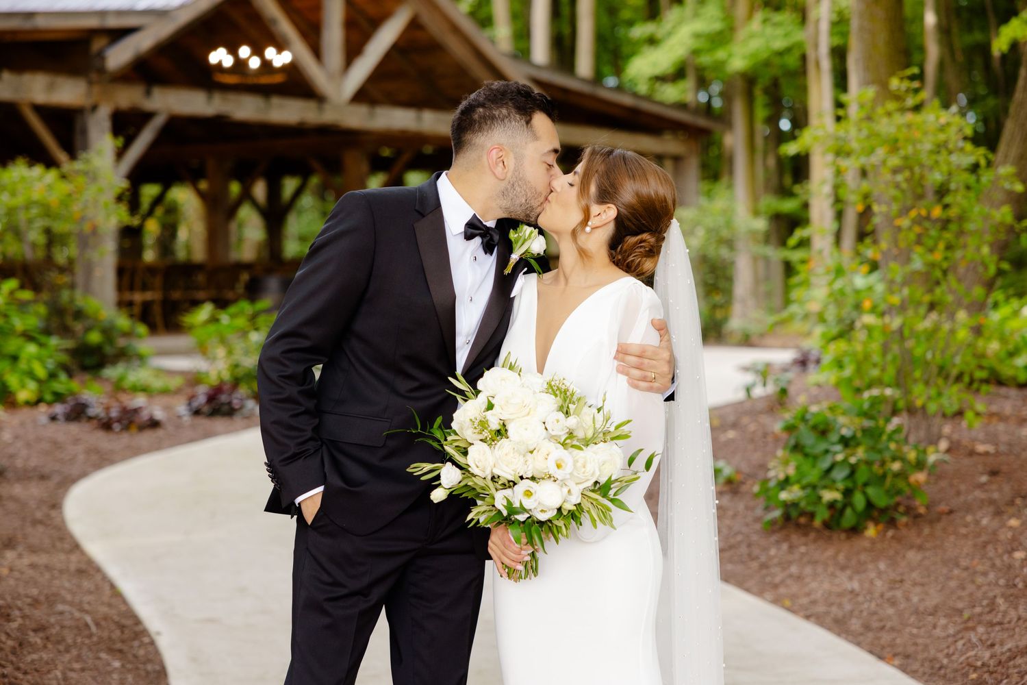 Romantic wedding portrait of couple on curved garden path with rustic woodland pavilion.