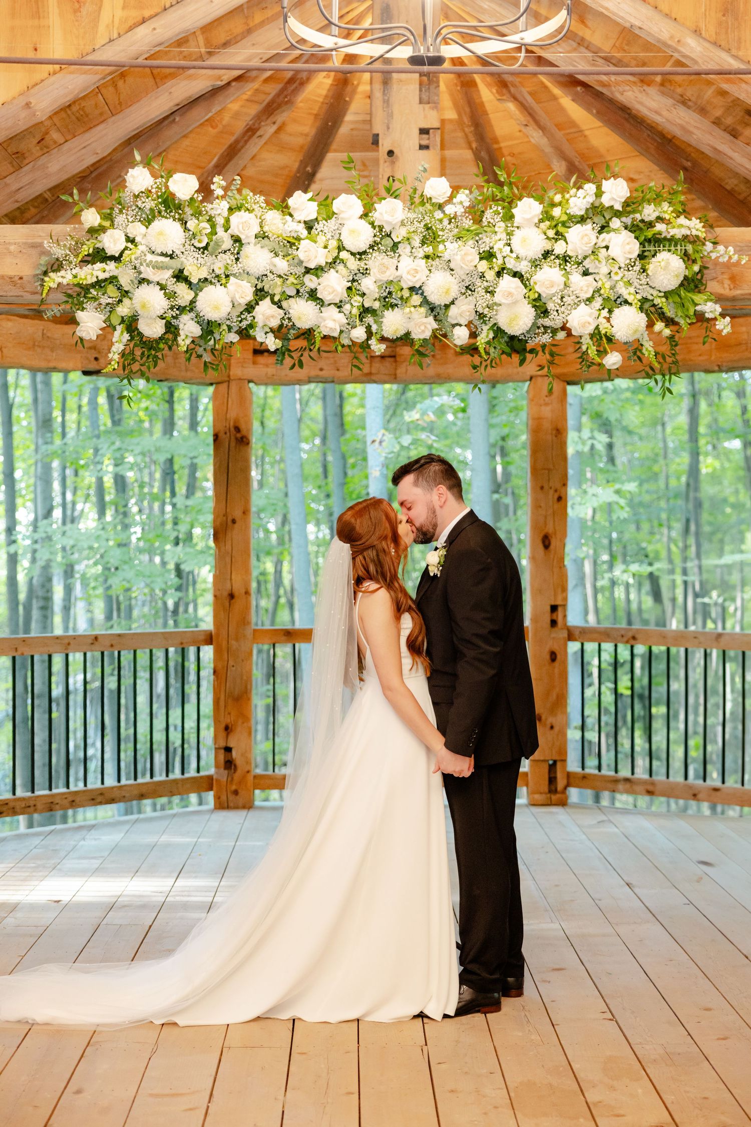 A wedding couple stands beneath a stunning white floral chandelier in a wooden gazebo surrounded by forest.