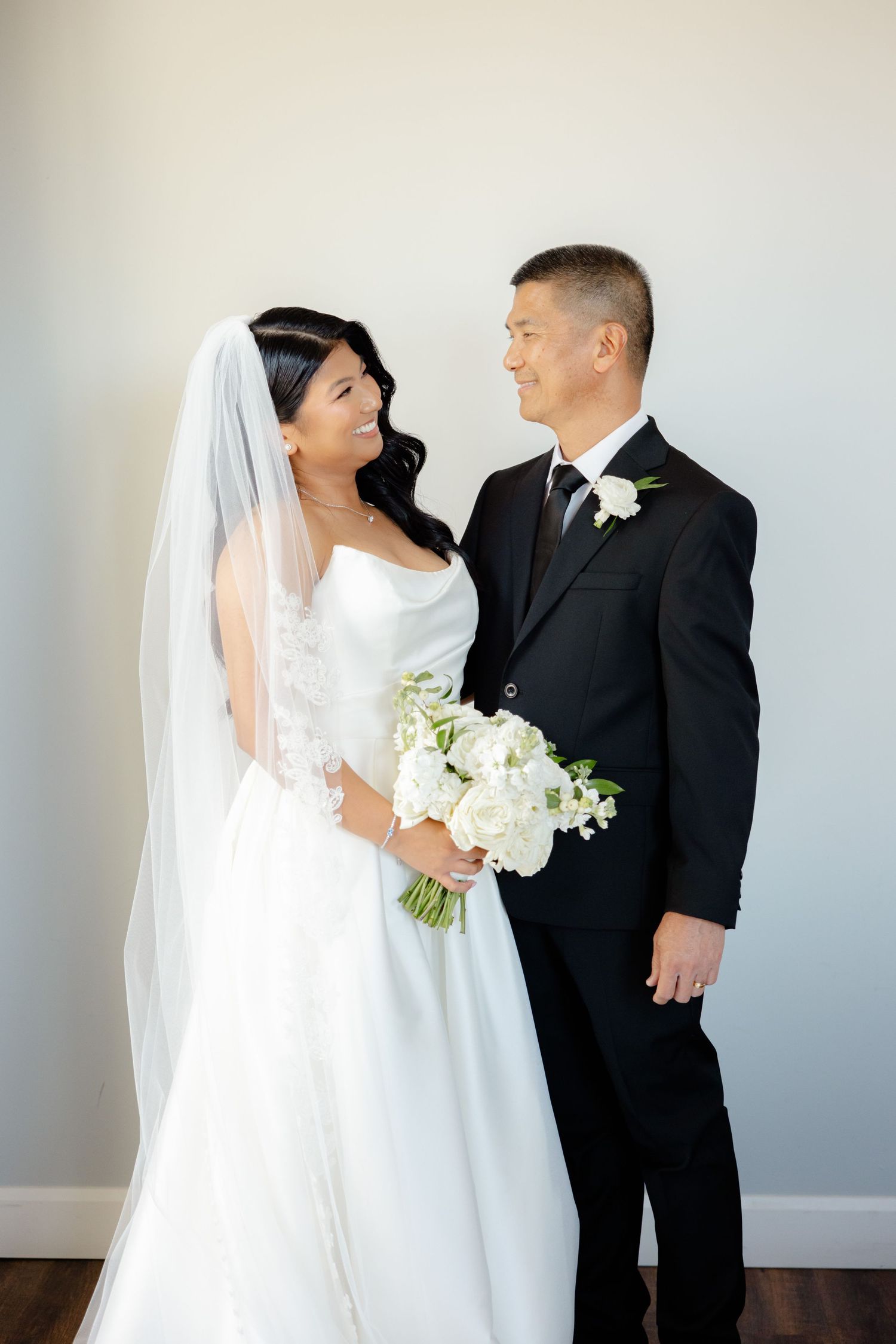 Bride in white dress and groom in black tuxedo pose together while holding a white floral bouquet against neutral wall.