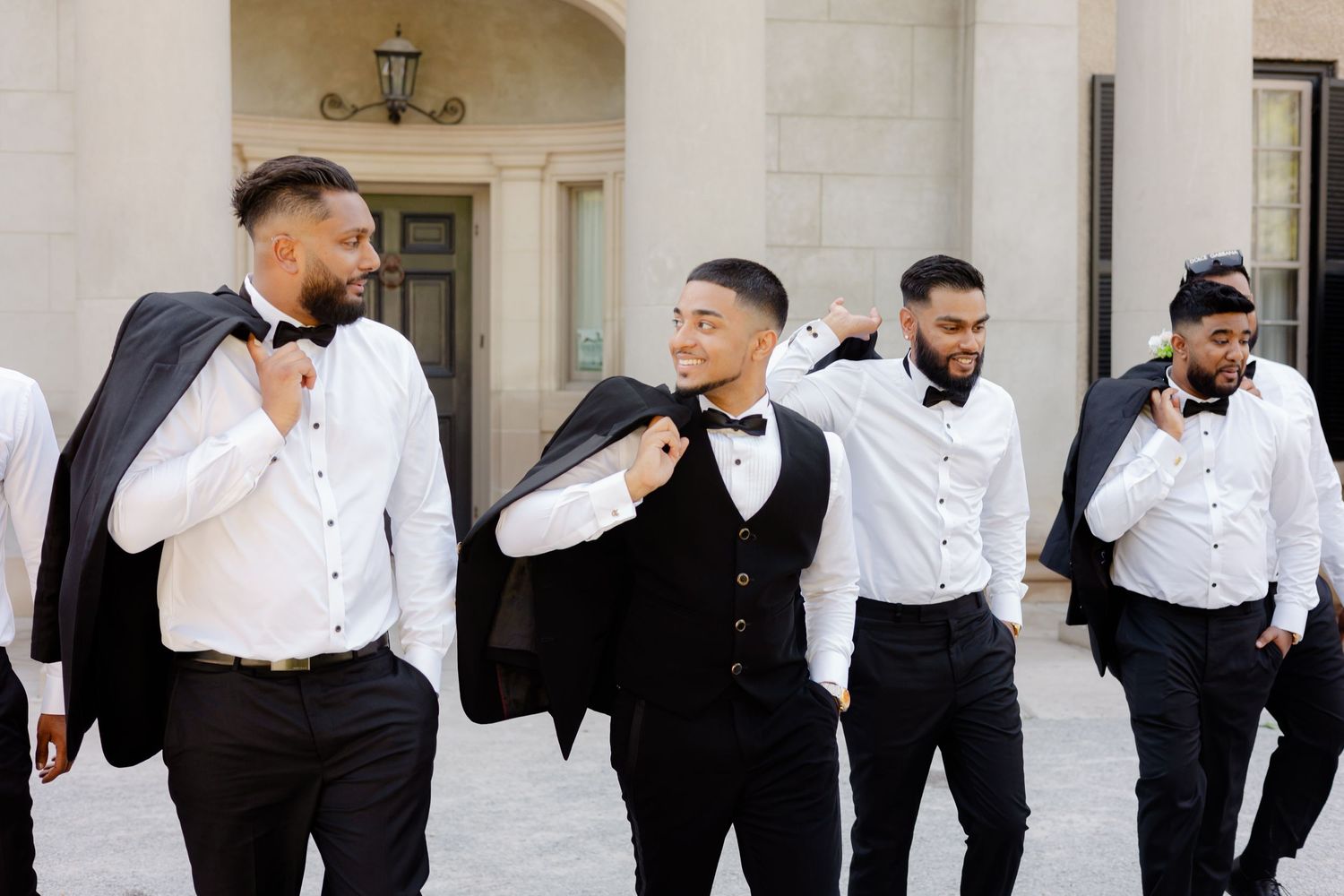 Groomsmen adjust their bowties in unison while standing outside a classical building entrance.
