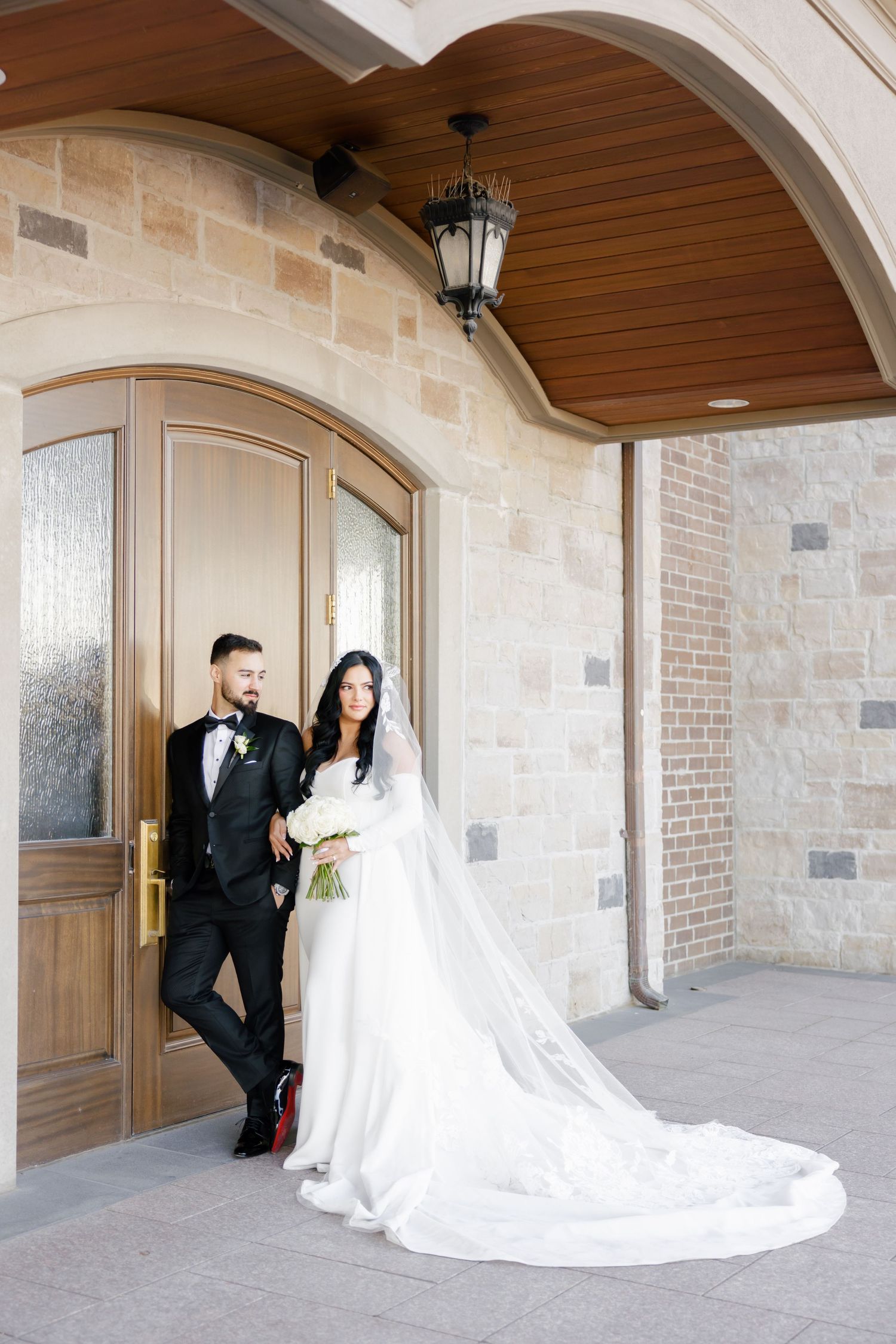 Wedding couple leans against stone wall with dramatic cathedral length veil trailing behind.