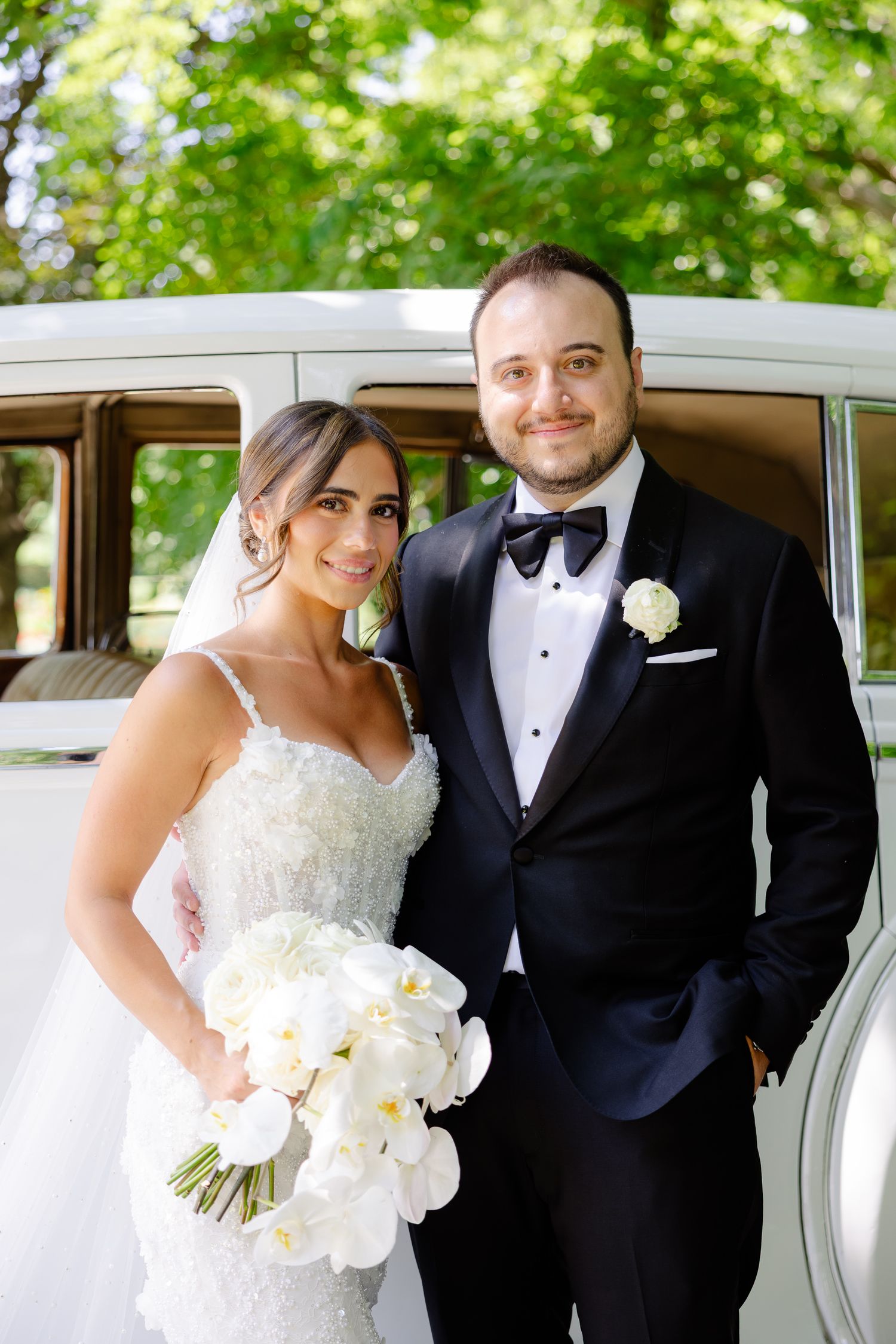 Wedding couple poses together in formal attire next to vintage white car during outdoor summer celebration.