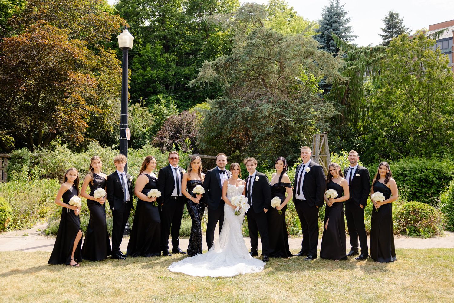 Wedding party in black formal attire poses on green lawn with trees and lamp post in background.