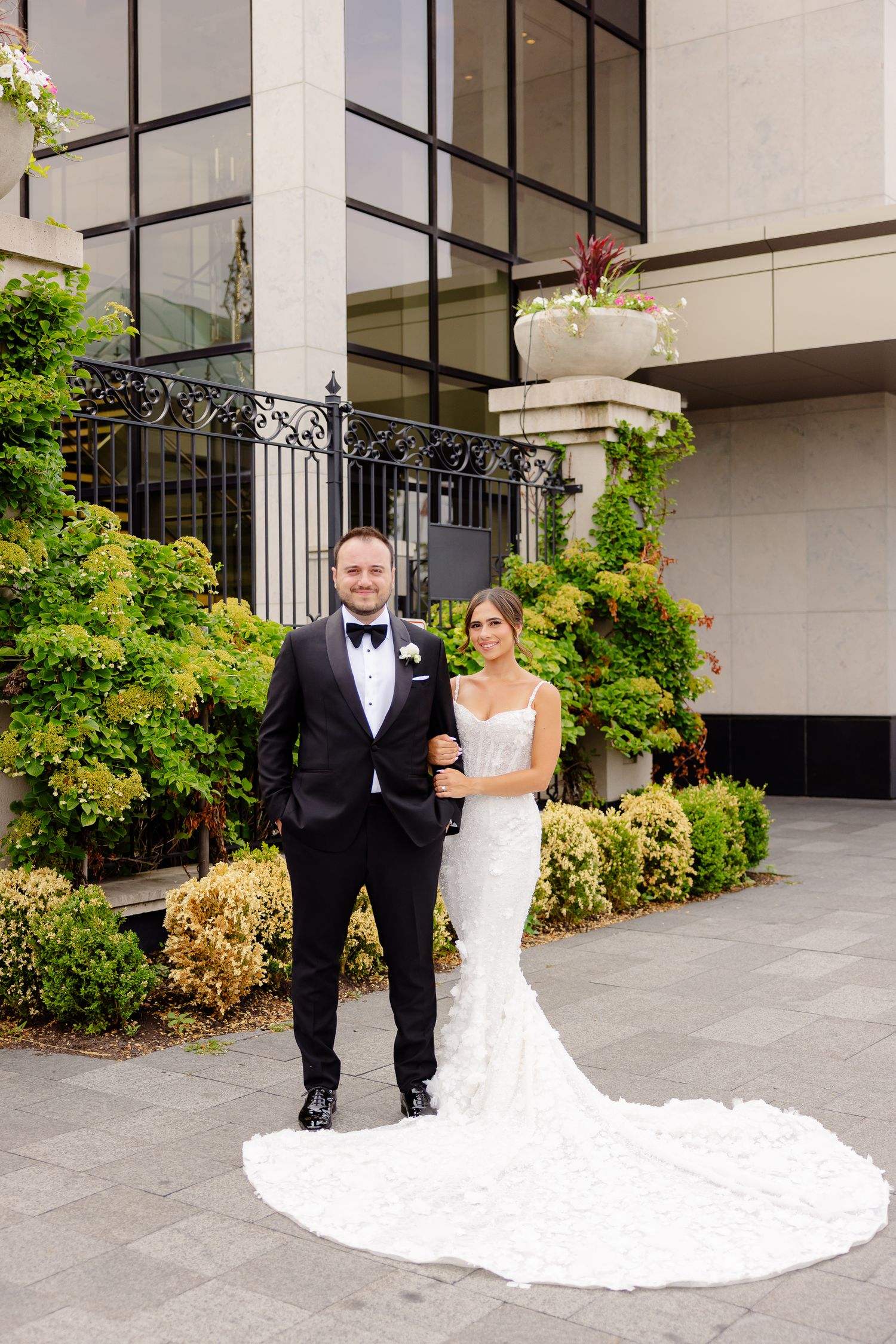 Elegant couple in formal wedding attire stand outside luxurious building with ornate railings and landscaping.