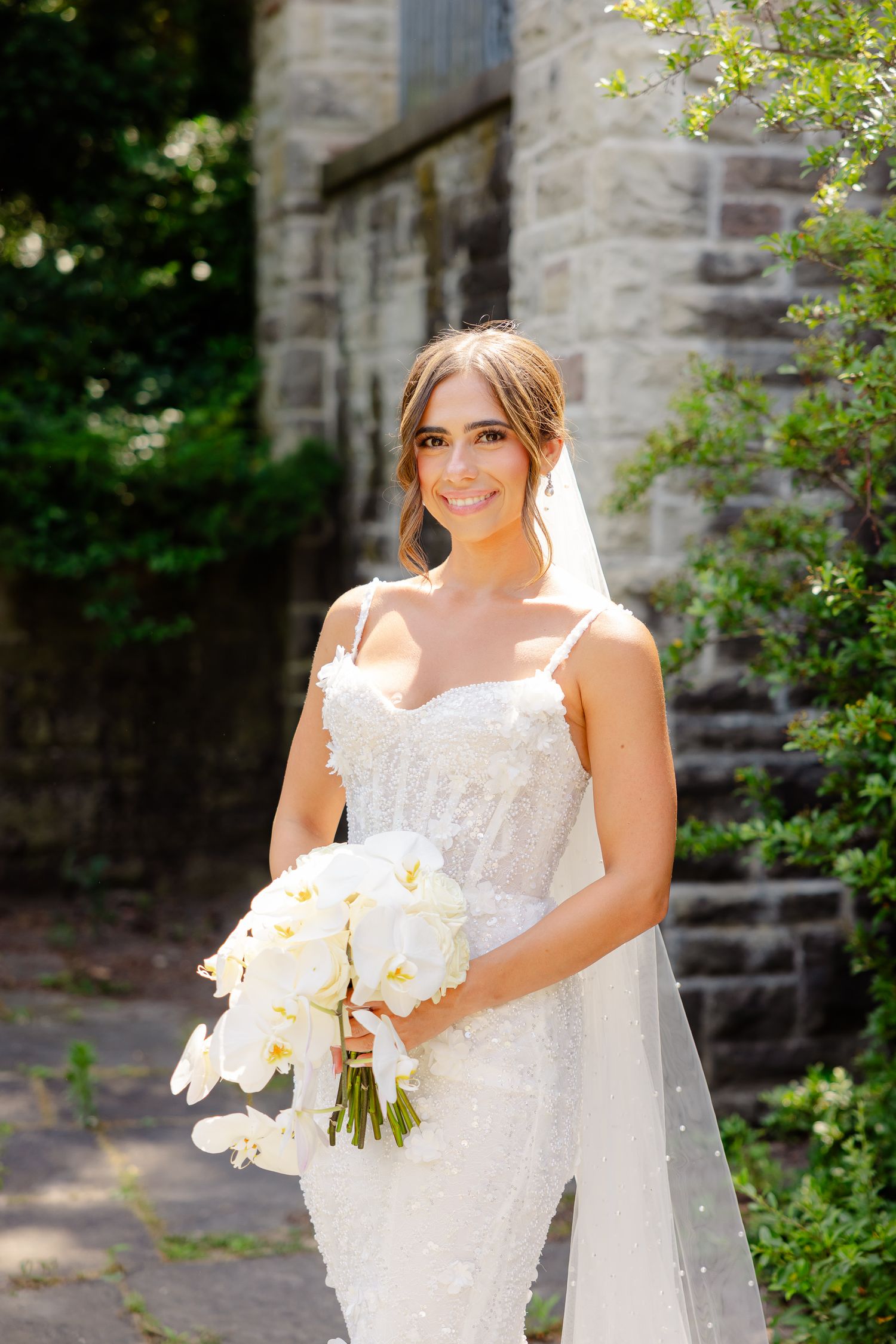 A bride in a fitted lace wedding gown holds a white orchid bouquet while standing beside a stone church wall.