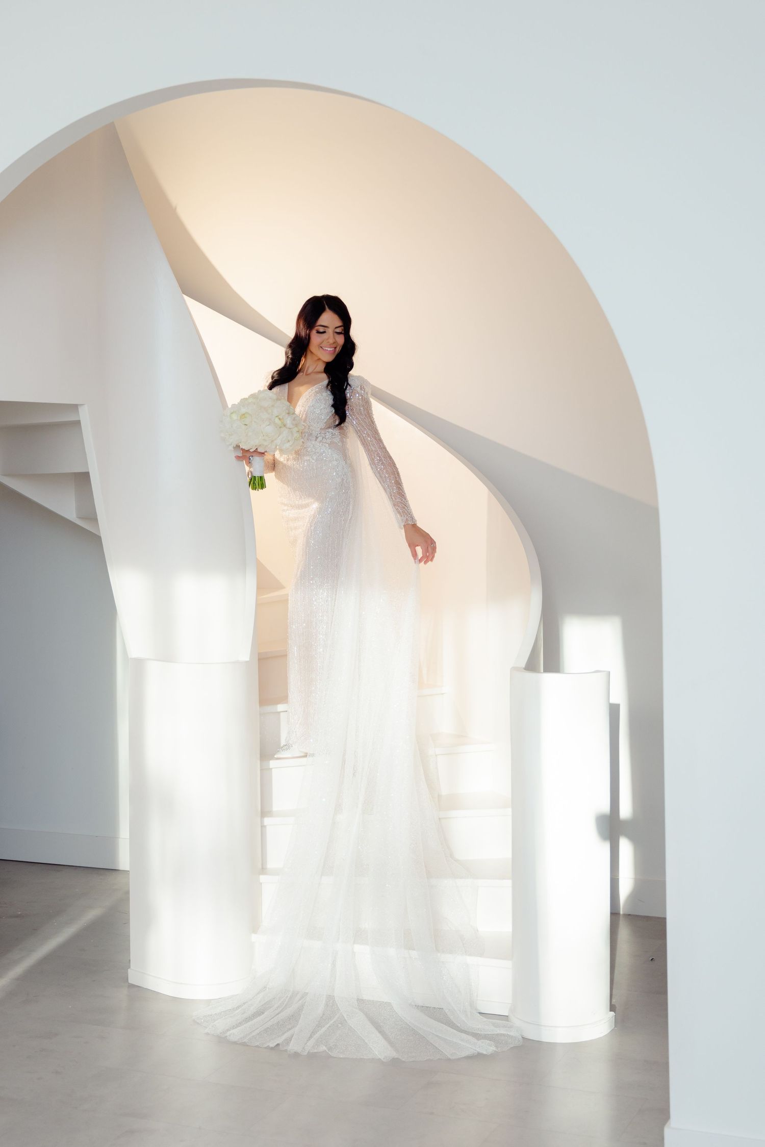 A flowing wedding dress train cascades down white steps under a minimalist curved archway.