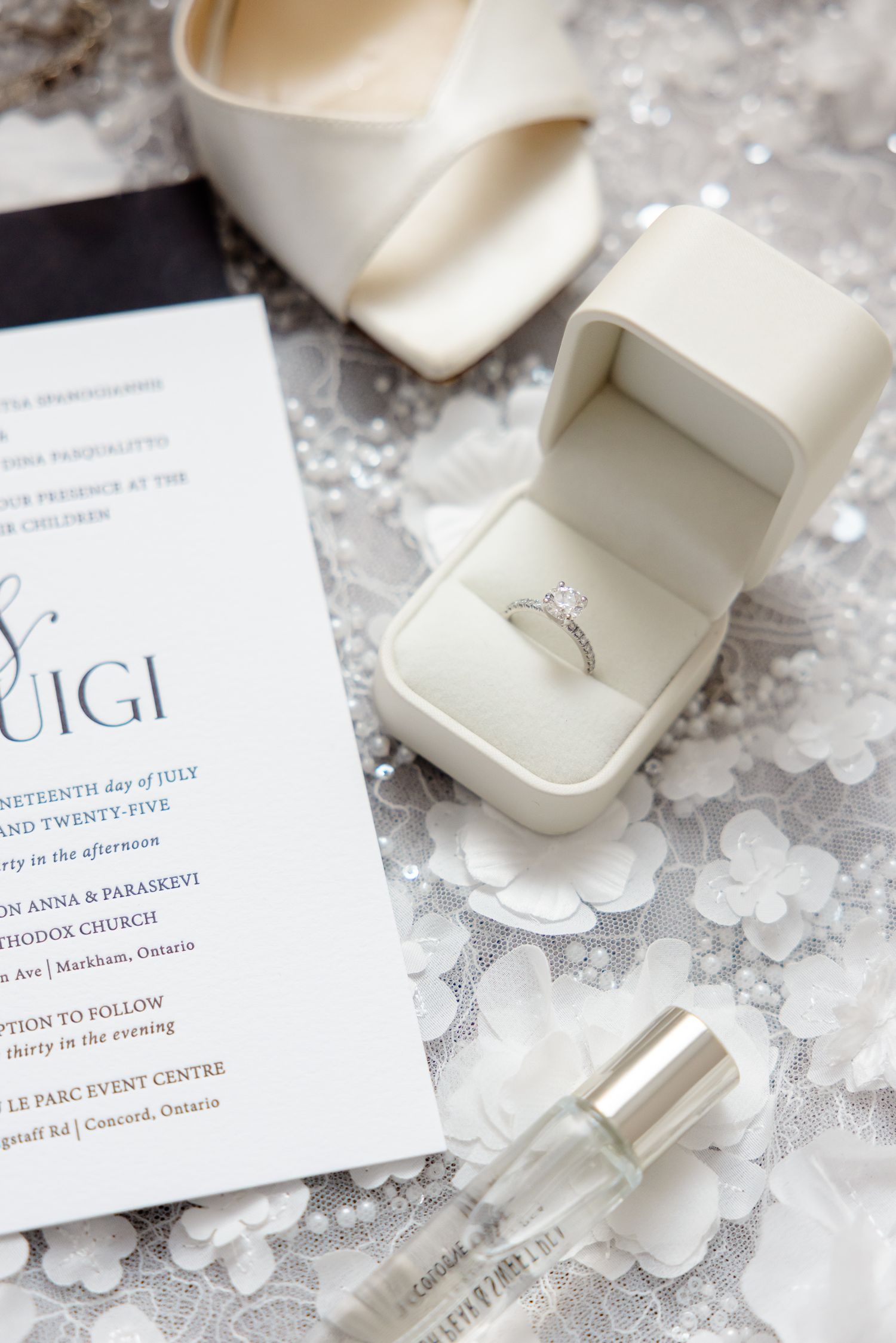 Wedding details with a white engagement ring box, invitation card, perfume bottle and ivory heels on a lace background.