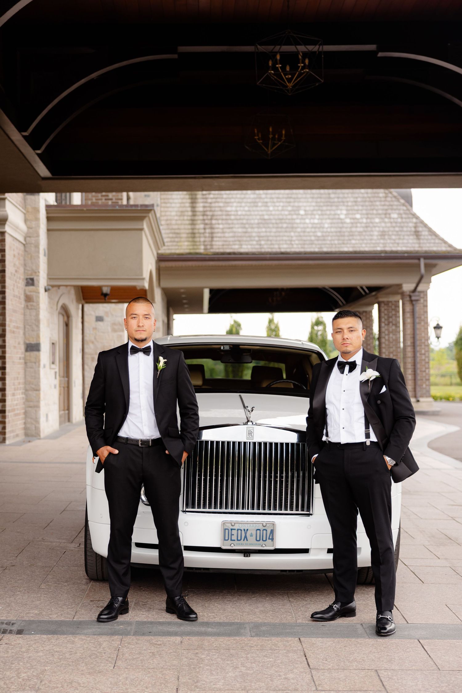 Two people in formal black tuxedos and bowties stand next to each other in front of a luxury vehicle outside a building.