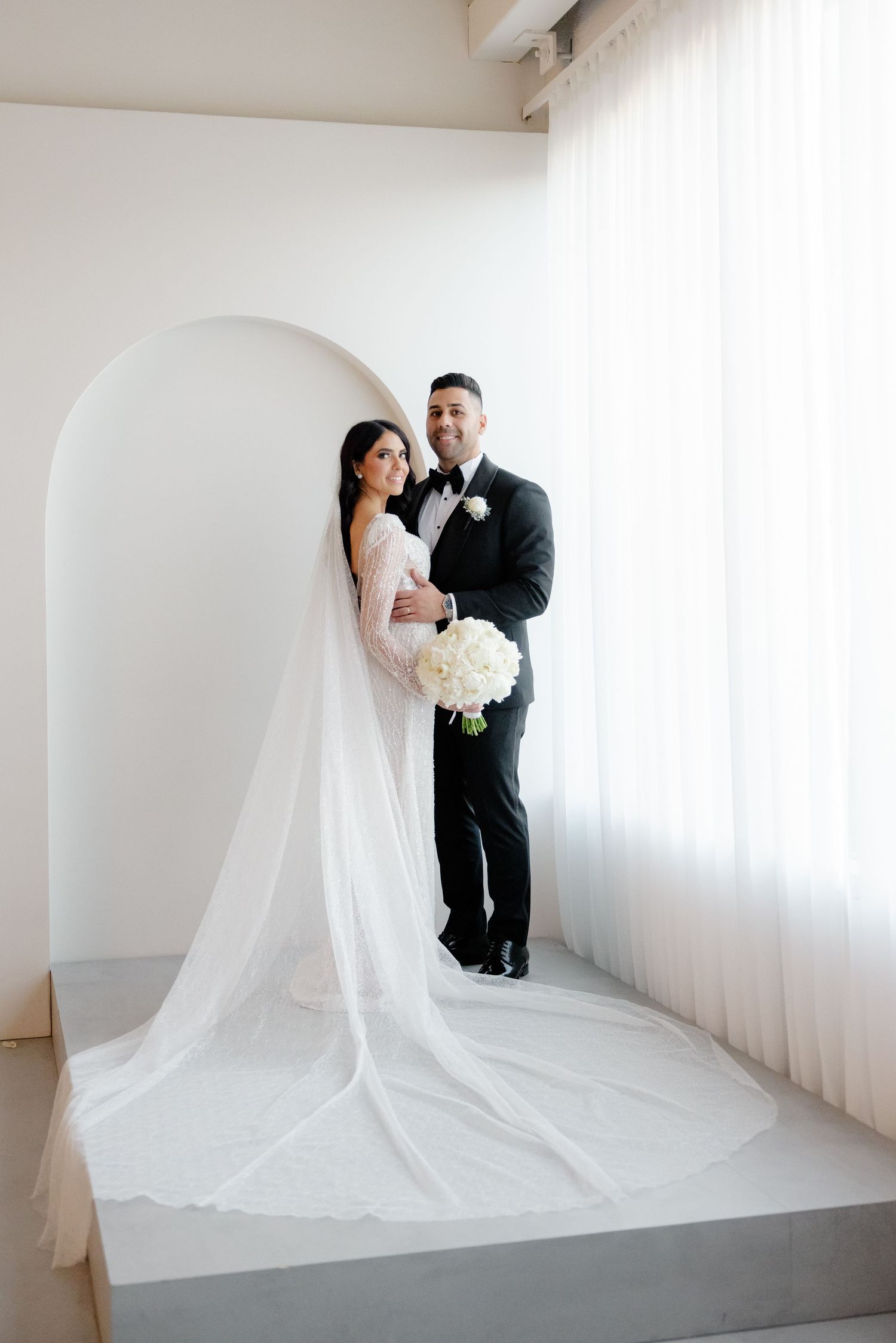 Newlywed couple stands in white archway holding bouquet with long veil trailing behind.