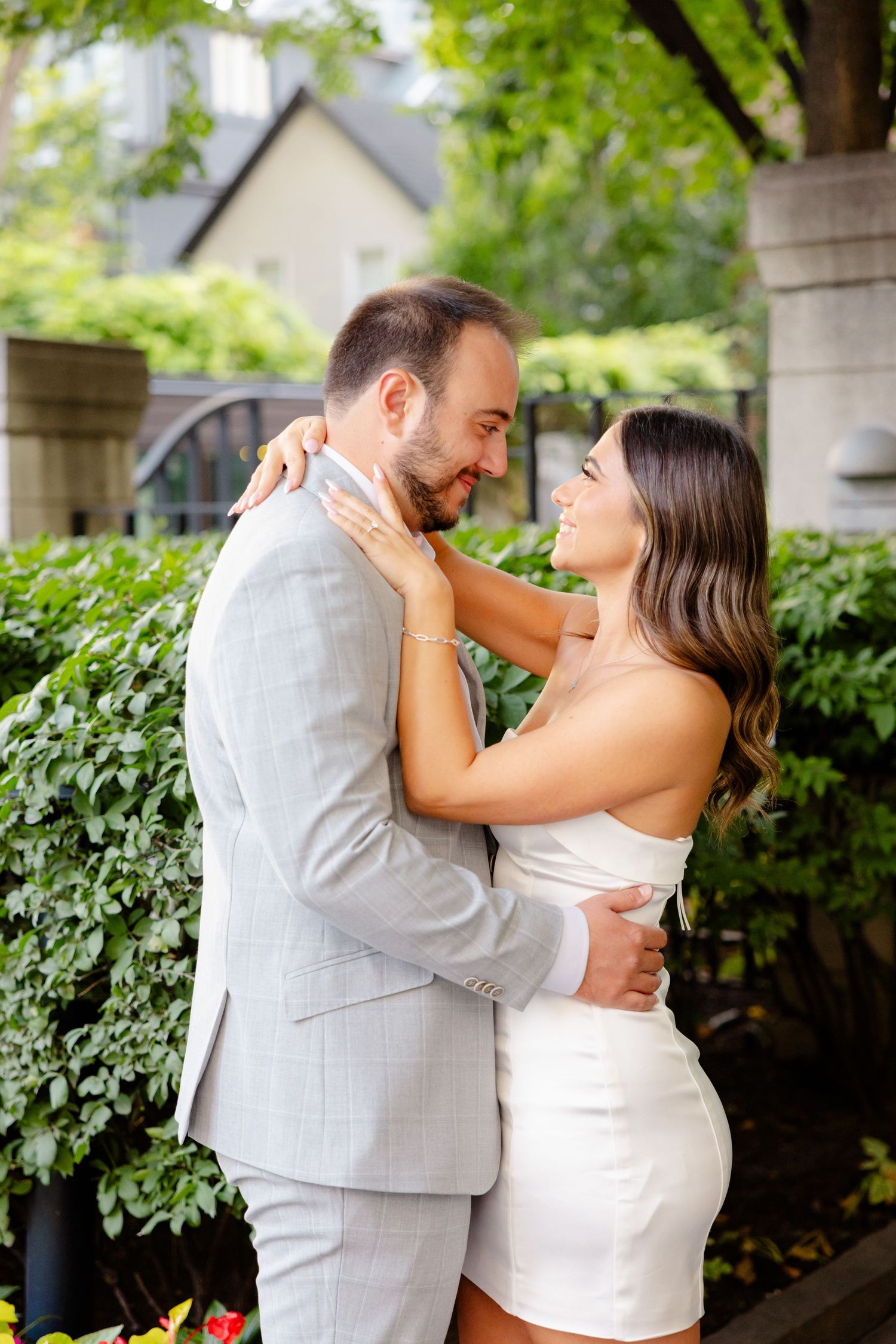 Elegant couple in white and grey attire embrace near garden wall with flowering plants.