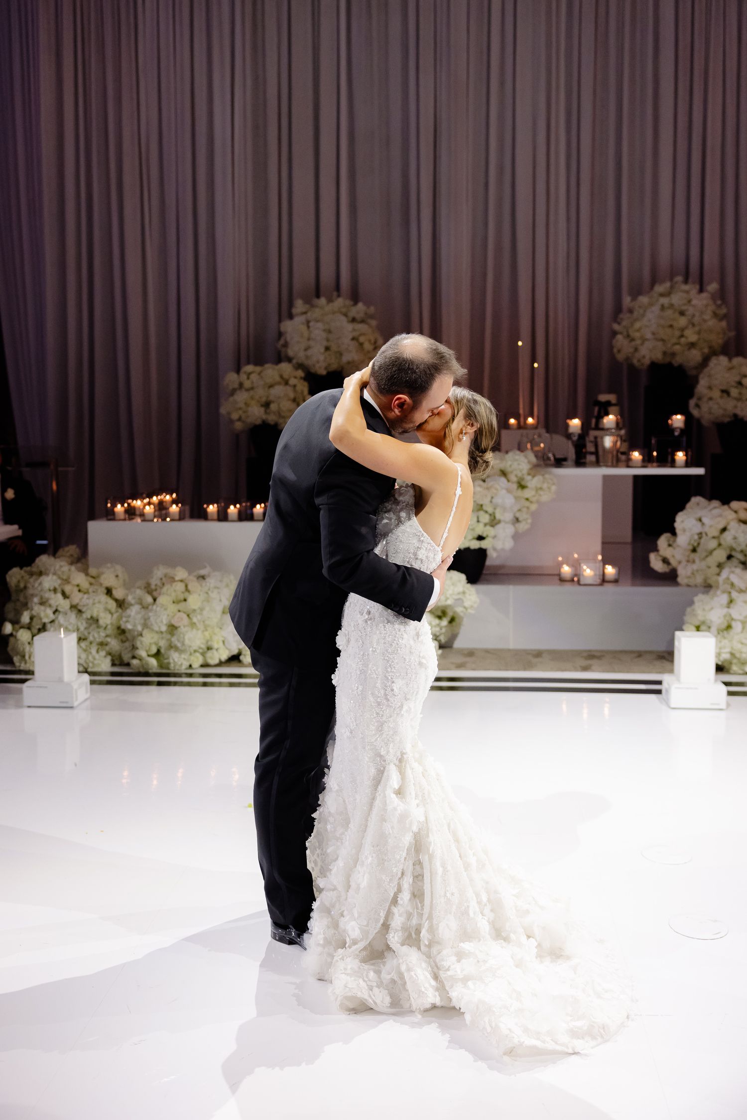 A romantic first dance between newlyweds on a white dance floor with floral arrangements and candlelight decor.