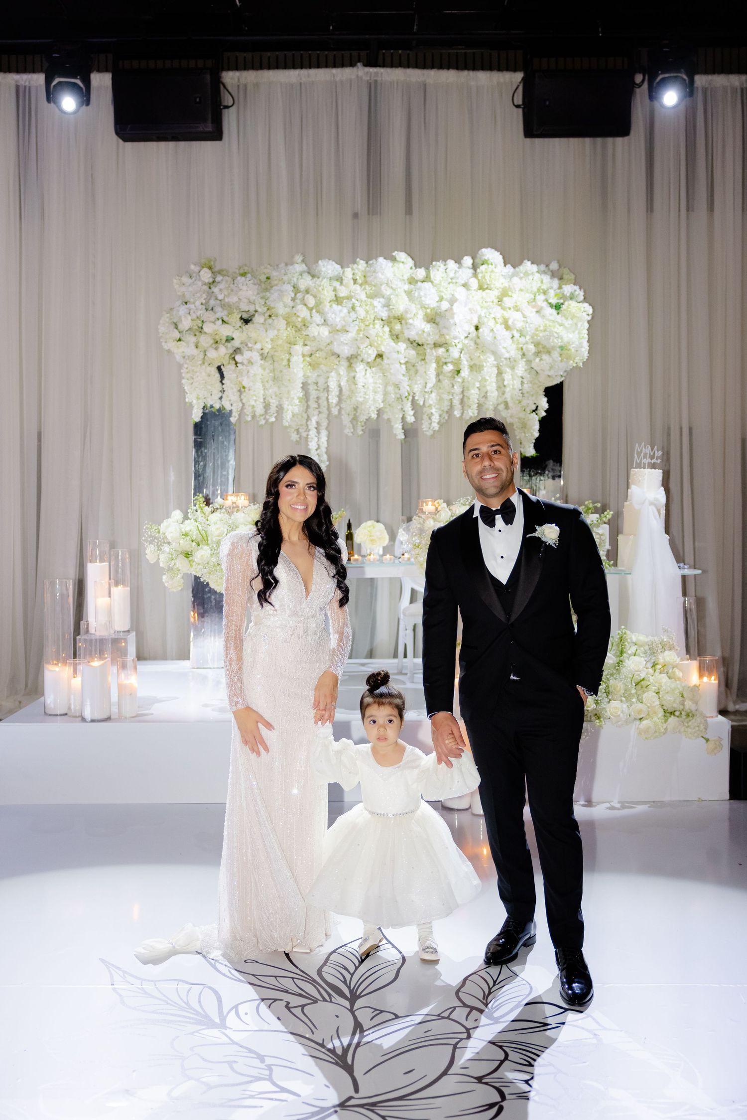 Wedding party poses beneath a stunning white floral chandelier on a decorated white stage.