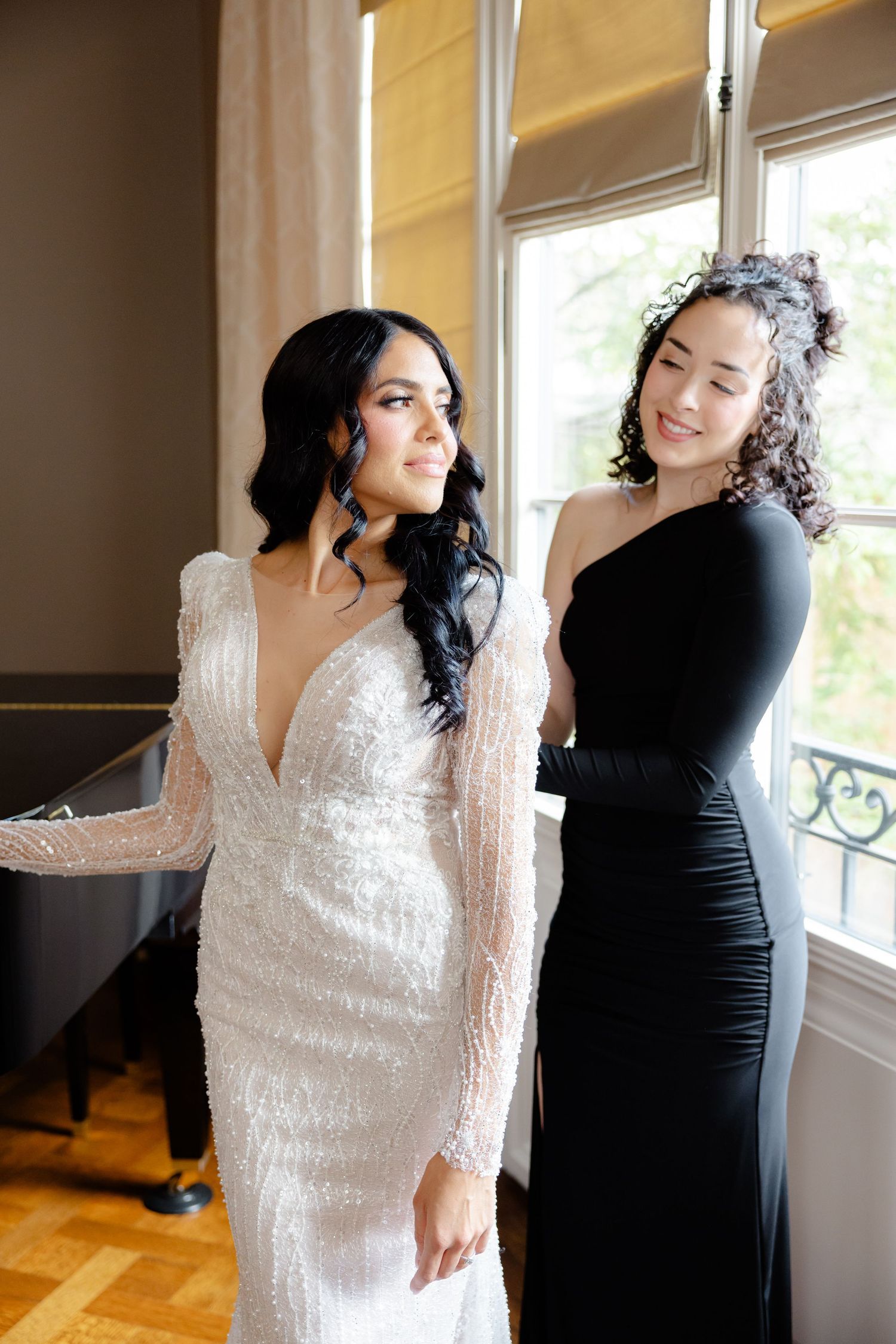 A bride in a sparkling white long-sleeve gown stands with someone wearing a black dress by a bright window.