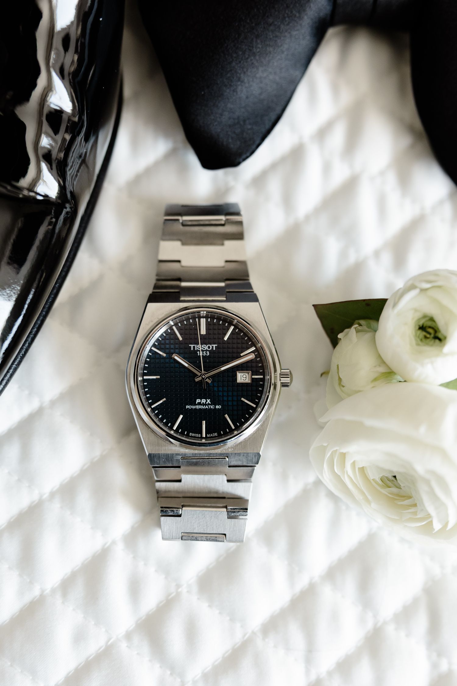 Elegant silver watch with black dial photographed on white fabric with delicate white flowers.