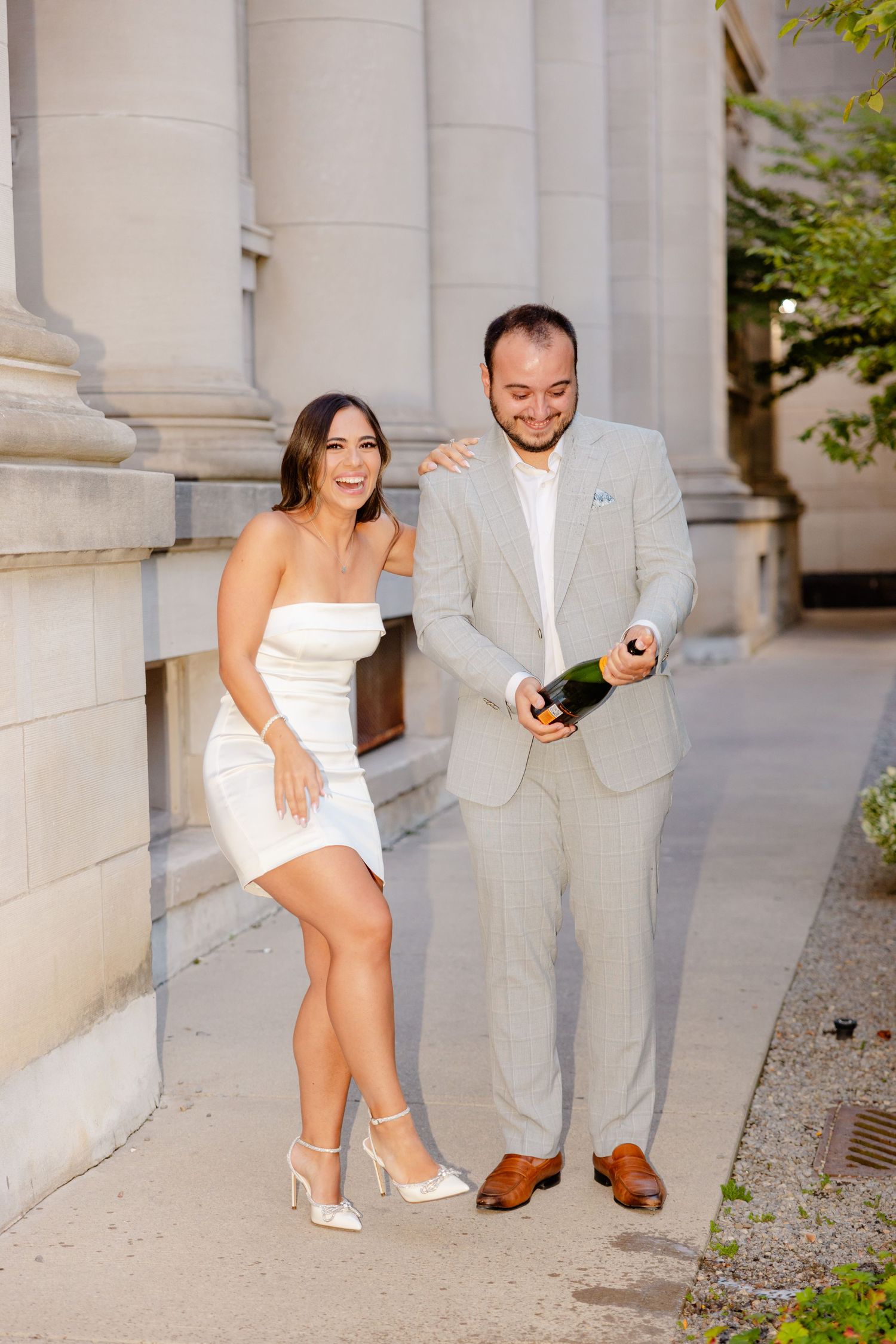 A couple in formal attire, with a white dress and light grey suit, poses together with champagne by stone columns.