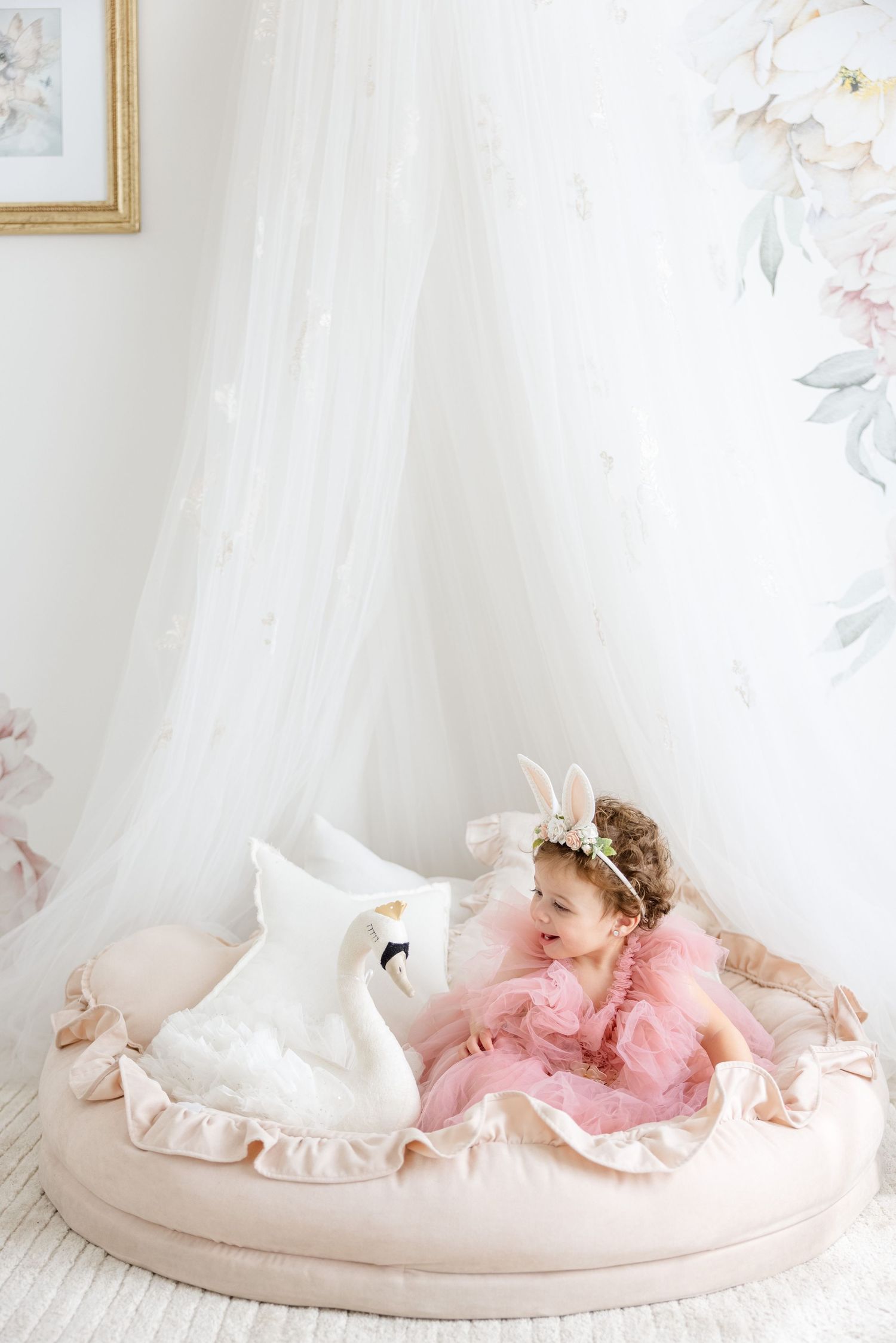 A toddler in pink tulle dress sits with white bunny decoration on a round beige cushioned platform.