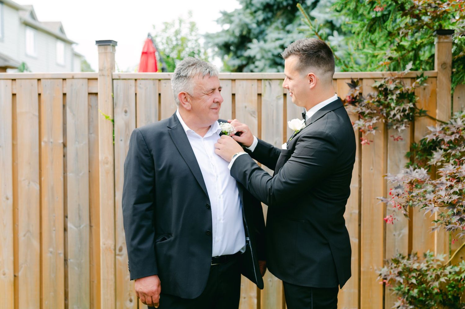 Two people share a moment while adjusting formal attire in front of a wooden fence.