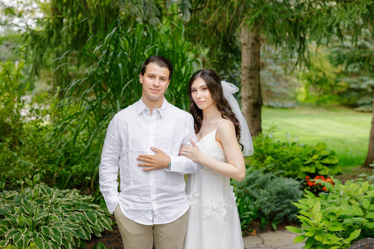 A wedding couple poses in a lush garden landscape wearing crisp white attire.