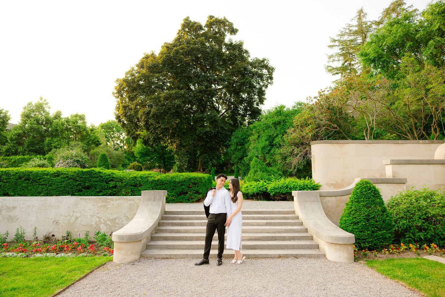 Couple stands together on curved stone steps in a formal garden with perfectly maintained landscaping.