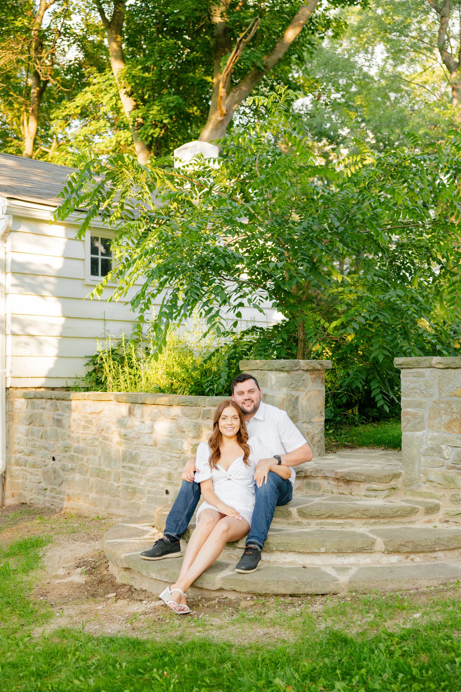 A couple sits casually on outdoor stone steps near a white house on a sunny day.