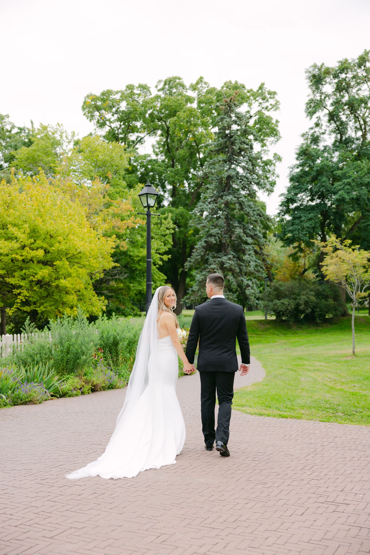 Wedding couple walks along brick pathway surrounded by lush gardens and tall trees.