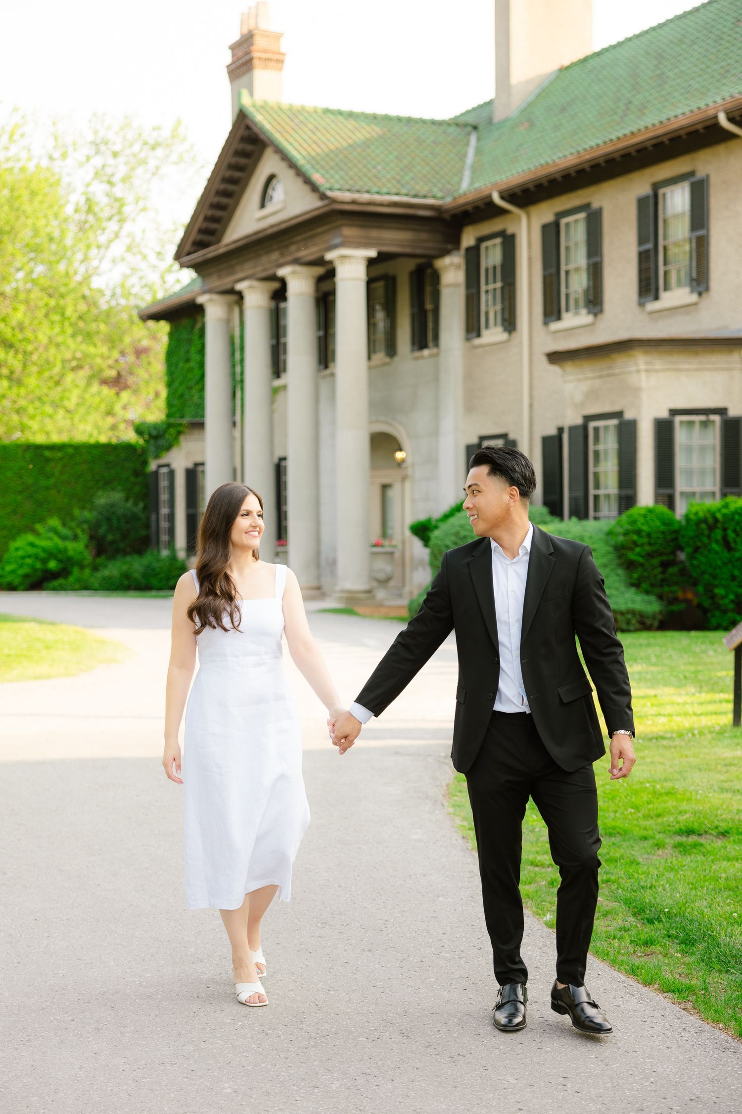 Couple holds hands while walking along path by grand colonial-style building.