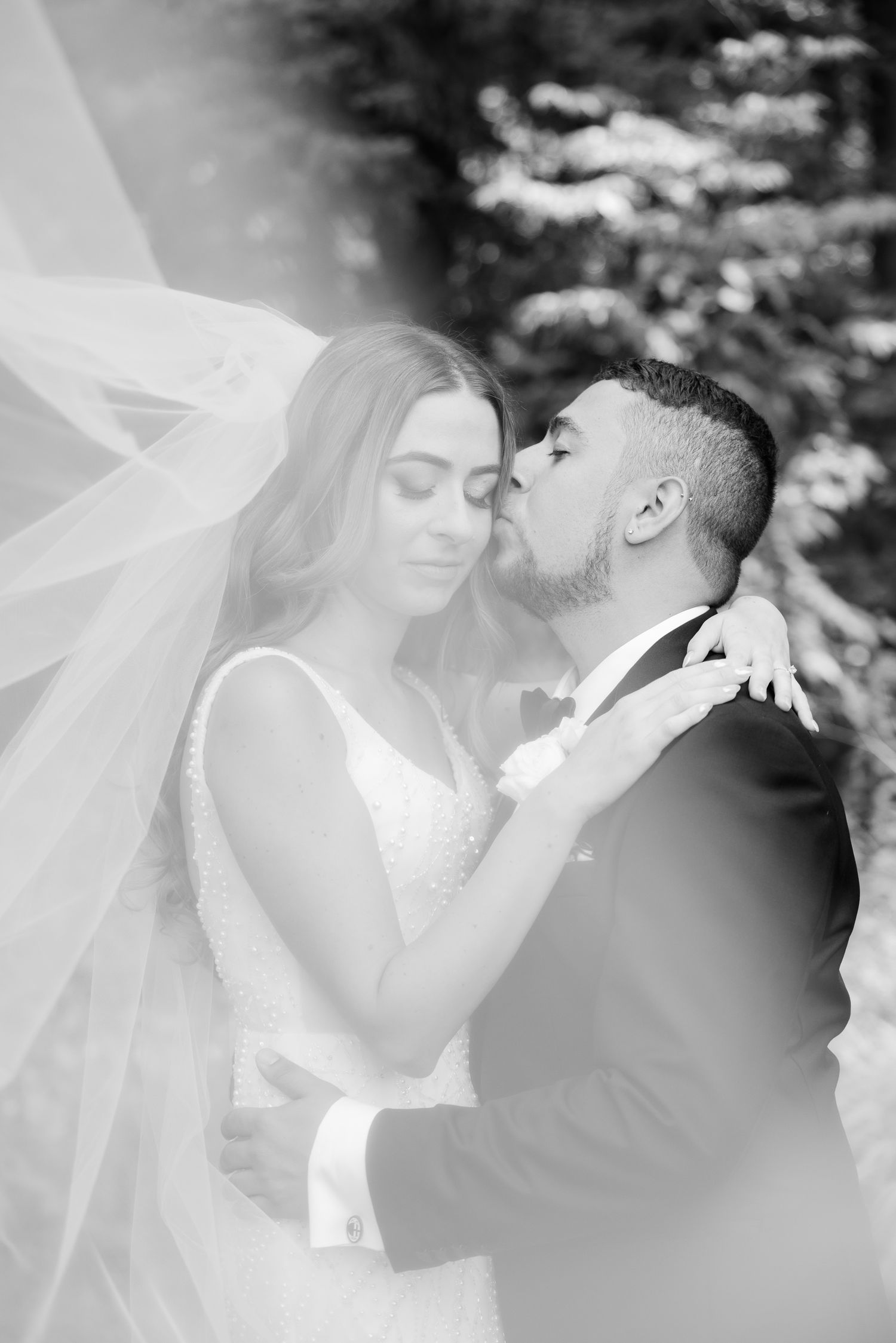 A romantic black and white wedding portrait showing a tender moment between newlyweds with flowing veil in natural setting.