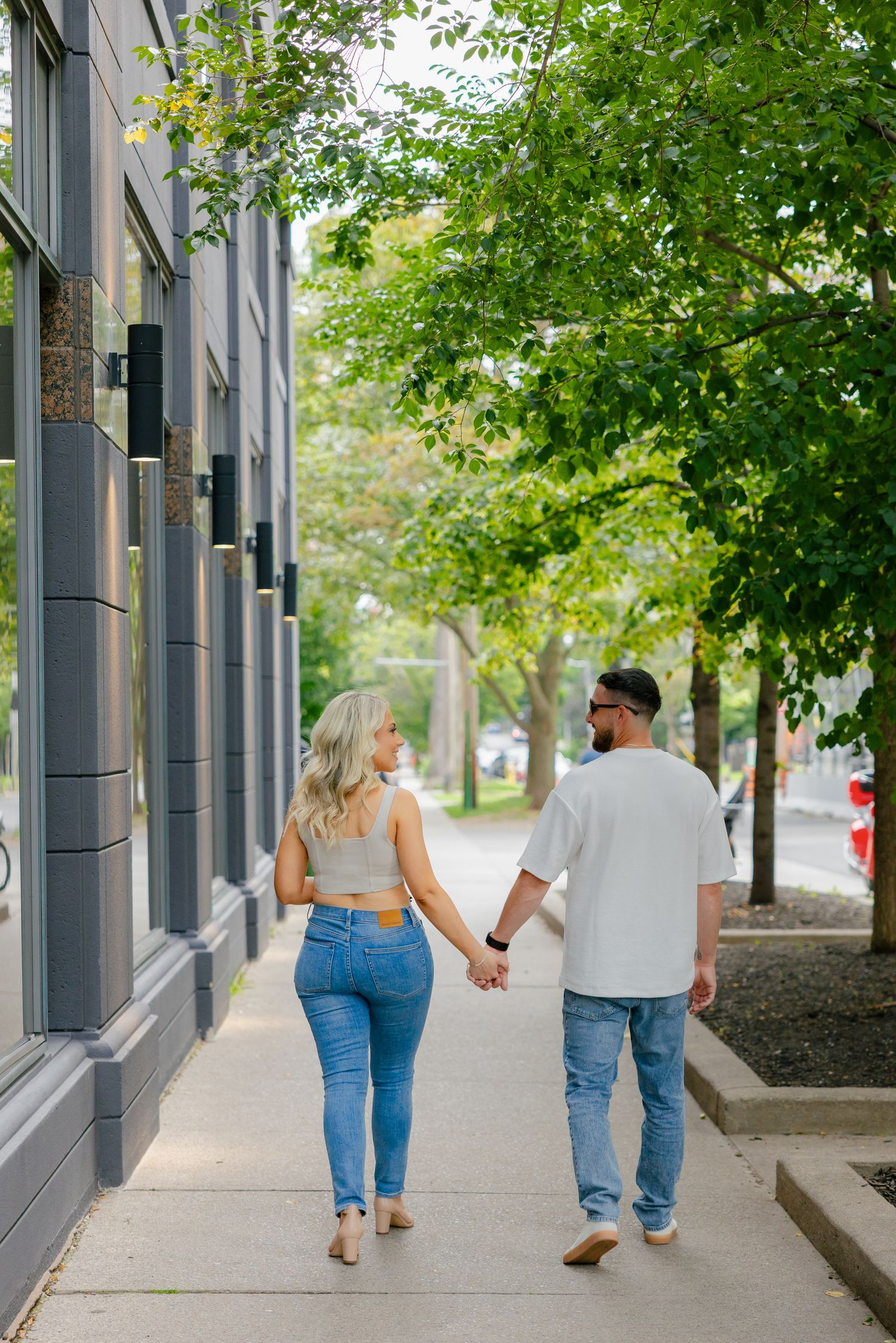 A couple walking hand in hand down a tree-lined urban sidewalk.