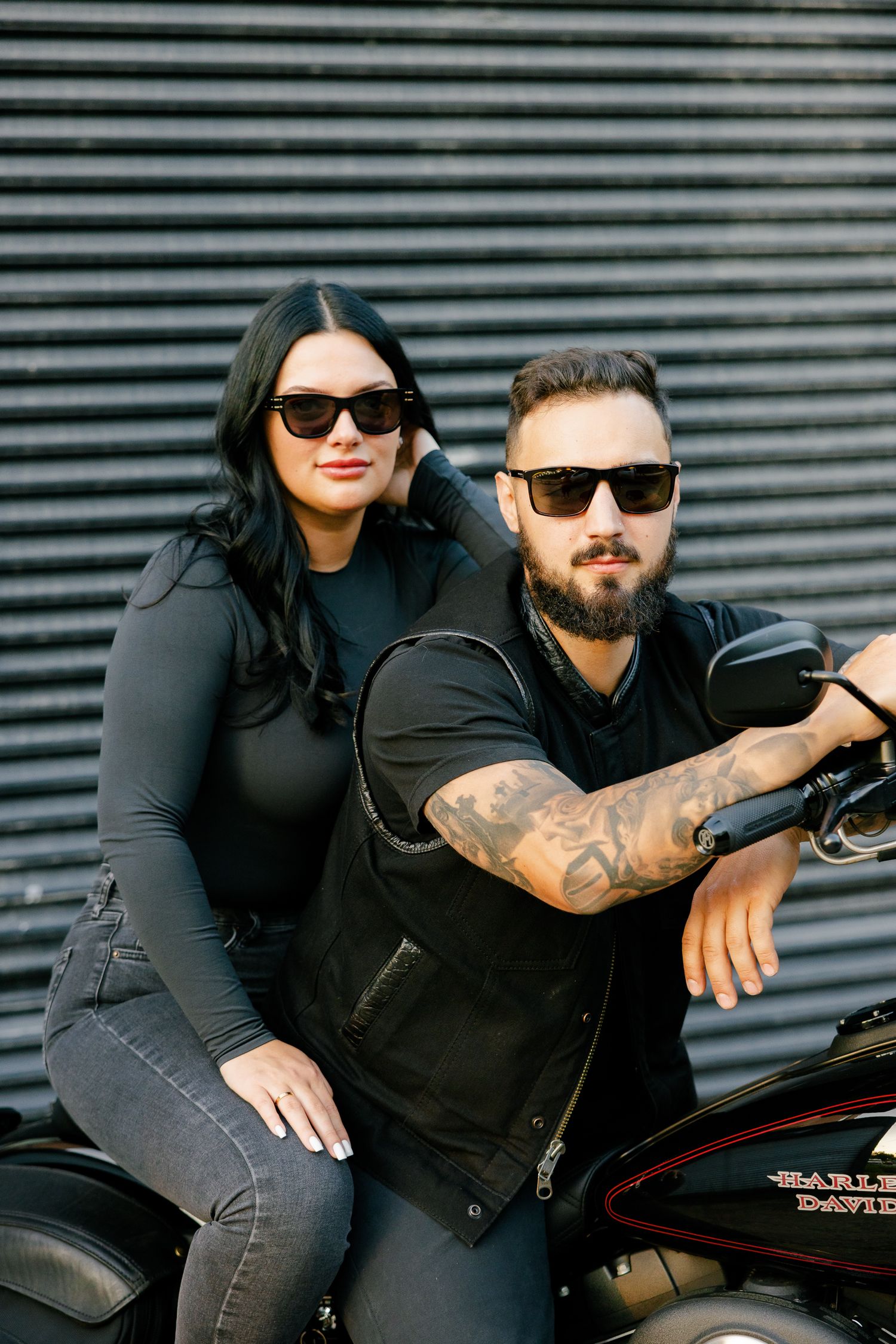 Stylish couple in black poses with Harley Davidson motorcycle against industrial striped wall.