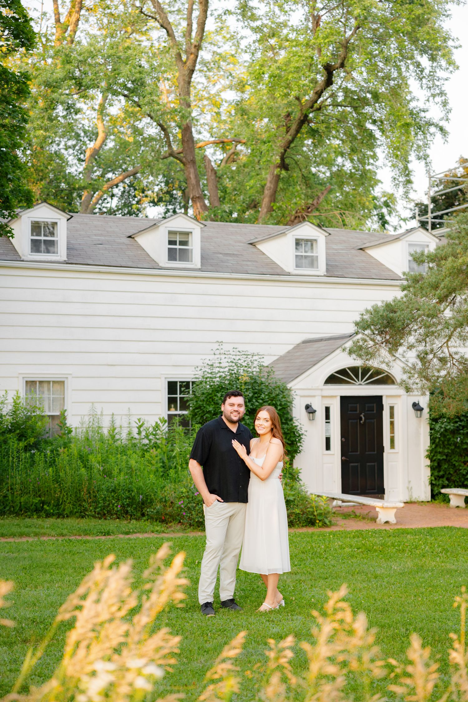 Summer portrait of couple in front of traditional white house with black door and dormers.