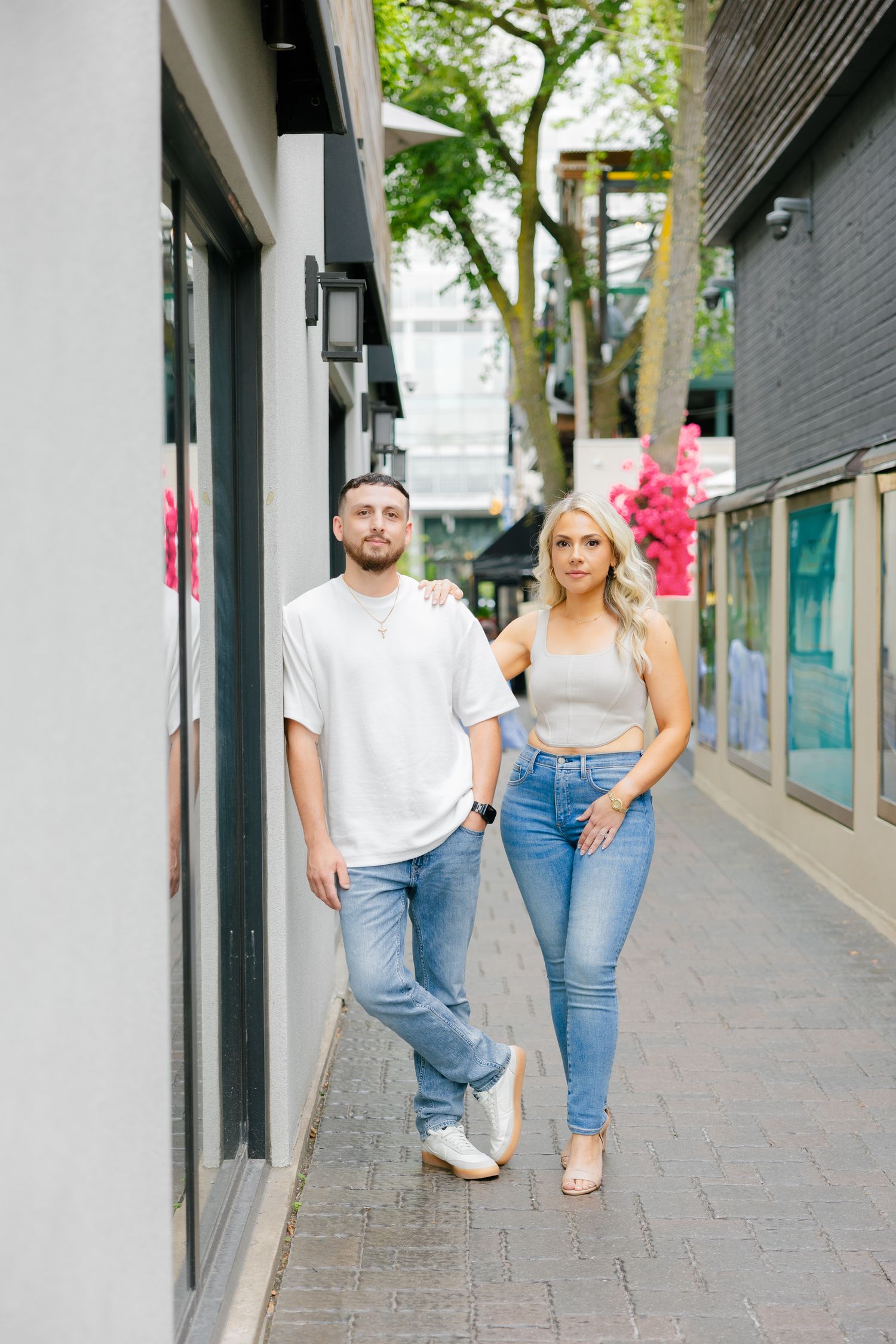 A casual couple in jeans and white tops posing in an urban alleyway with pink flowers.