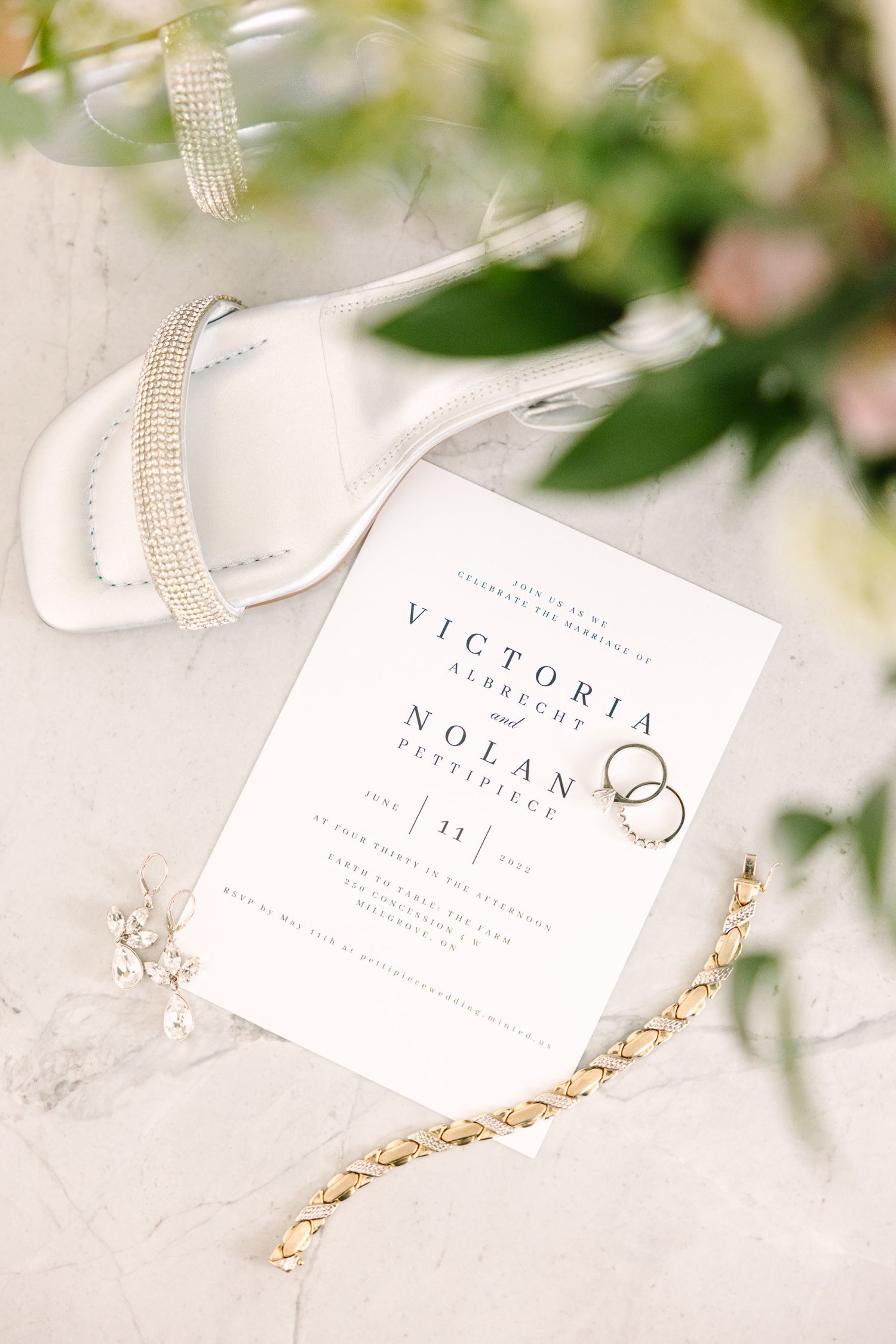 Wedding invitation with gold jewelry and white sandals styled with greenery.