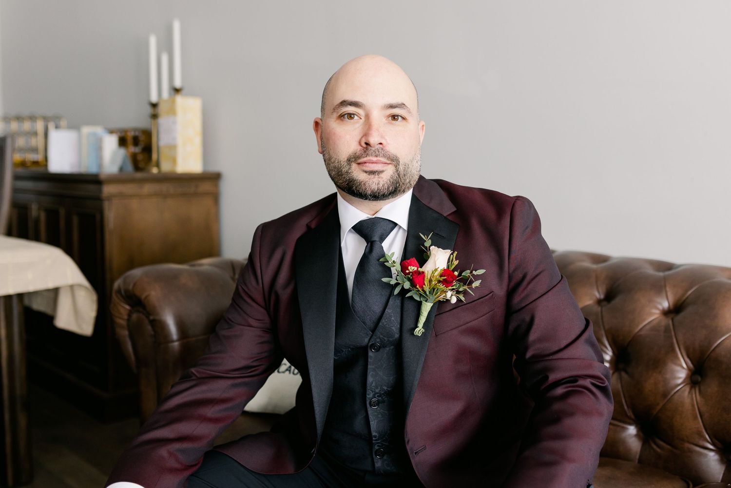 Groom in burgundy suit and black tie poses on leather couch with red floral boutonniere.