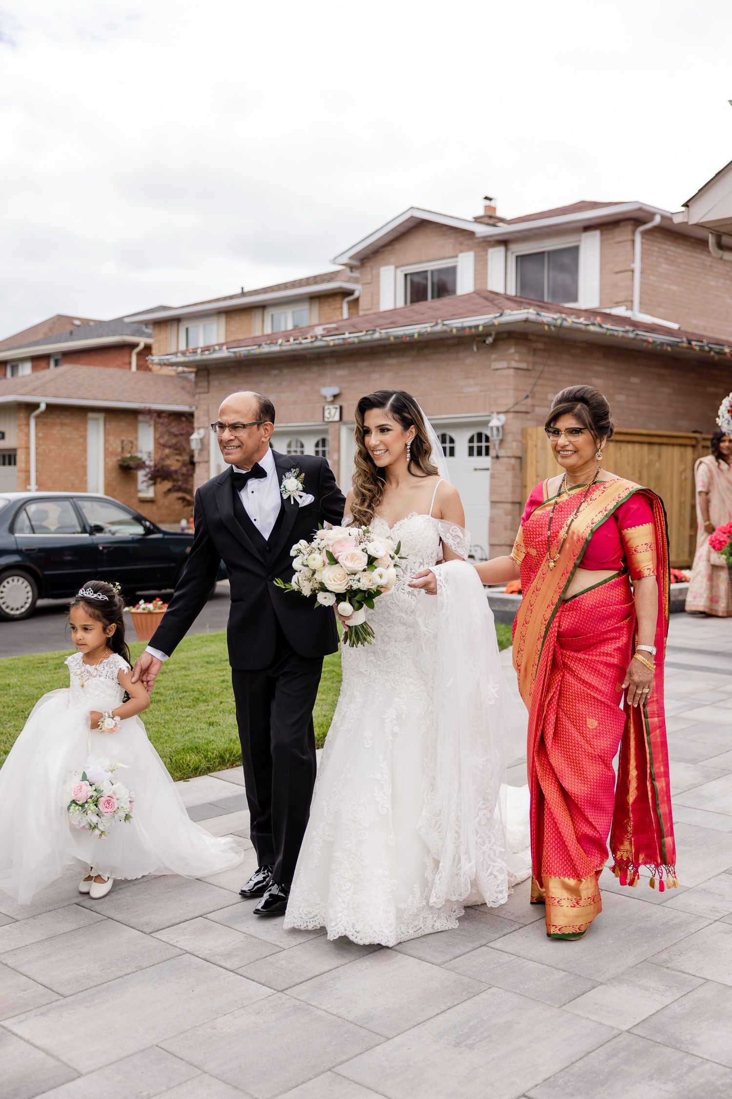 Wedding party walks together on stone pathway with bride in white gown and guest in vibrant red sari.